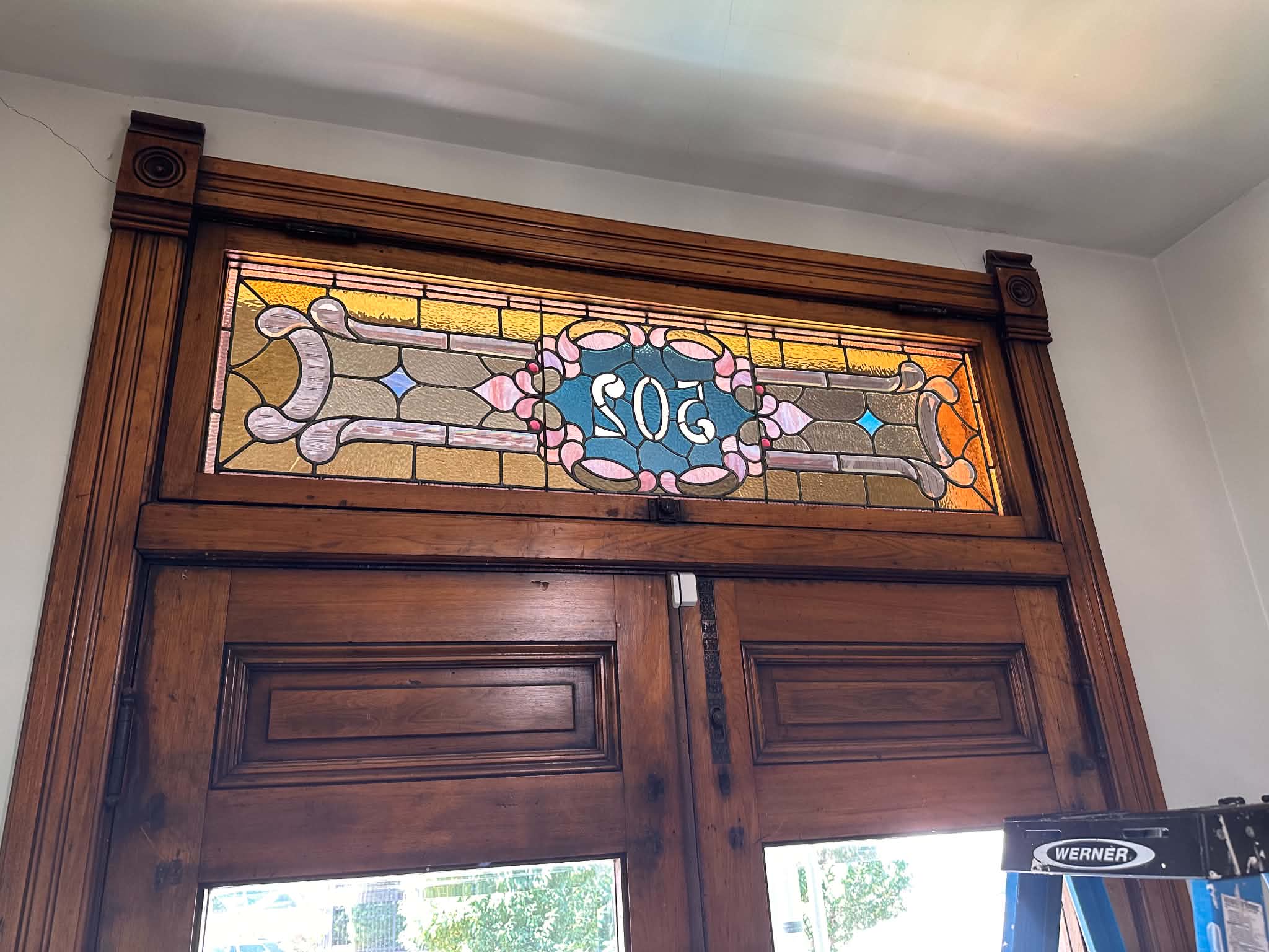 Decorative stained glass window with the number 502, framed by dark wood, above a wooden door, with a glimpse of outside visible.