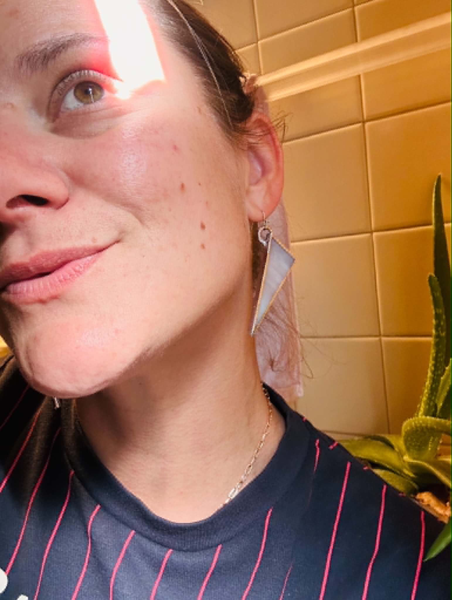Partial view of a smiling person's face with a yellow tiled wall and a plant in the background.