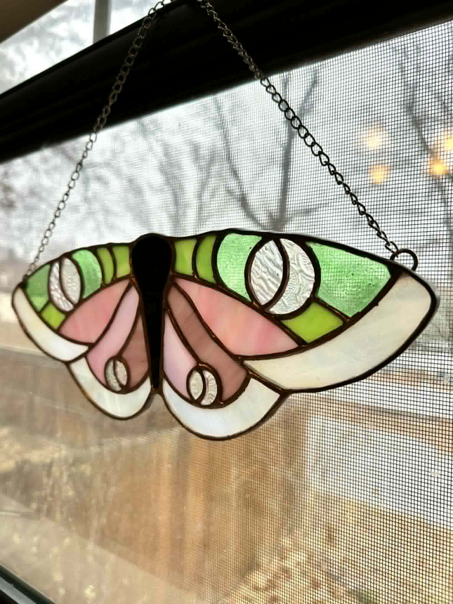 Colorful stained glass butterfly-shaped hanging decoration with green, pink, and white sections, hanging by chains in front of a window.