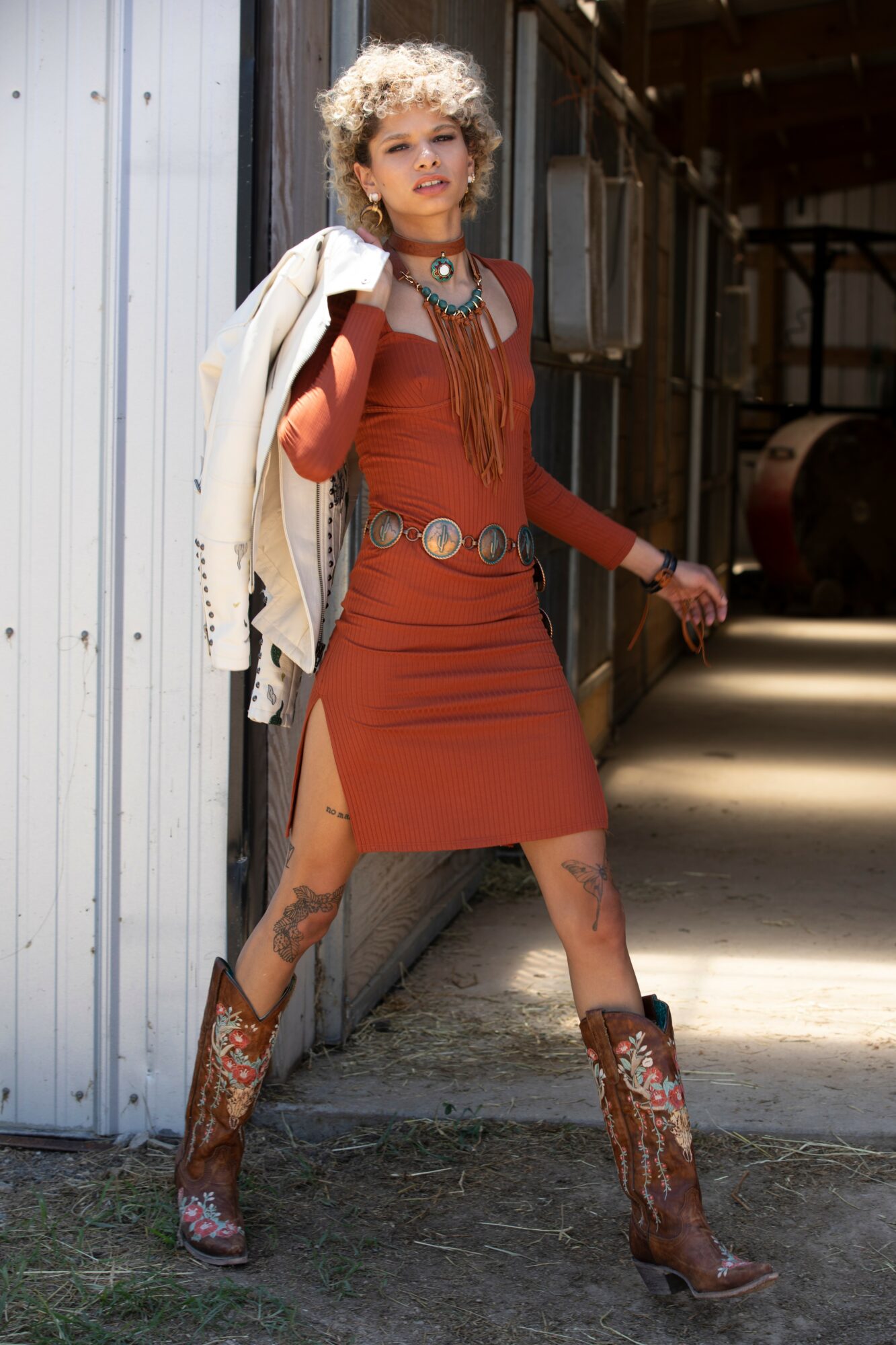 Woman with curly hair in a rust-colored dress, cowboy boots, and jewelry, standing outdoors near a building.