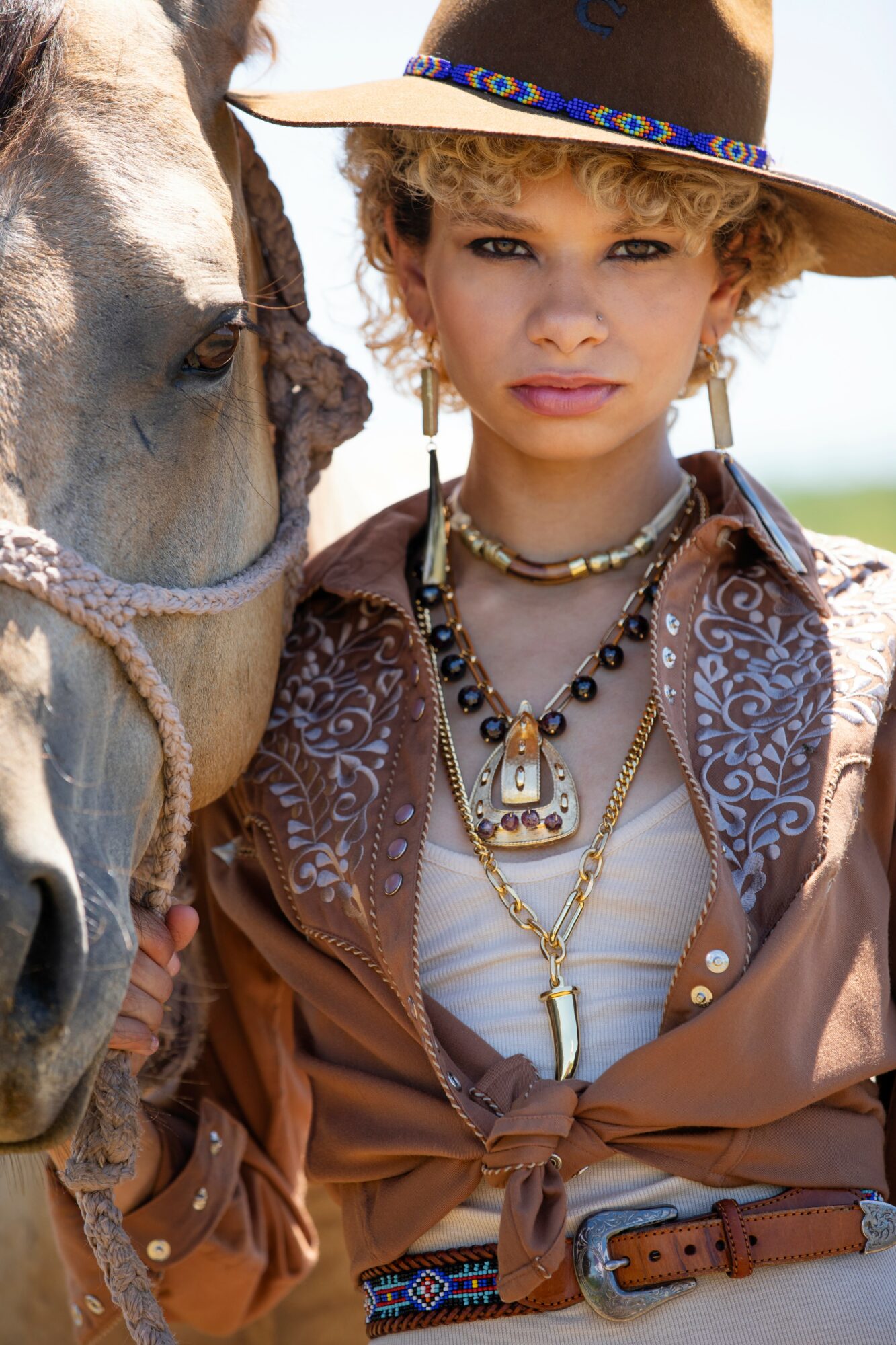 Young girl dressed in vintage clothing with jewelry, standing next to a horse outdoors.