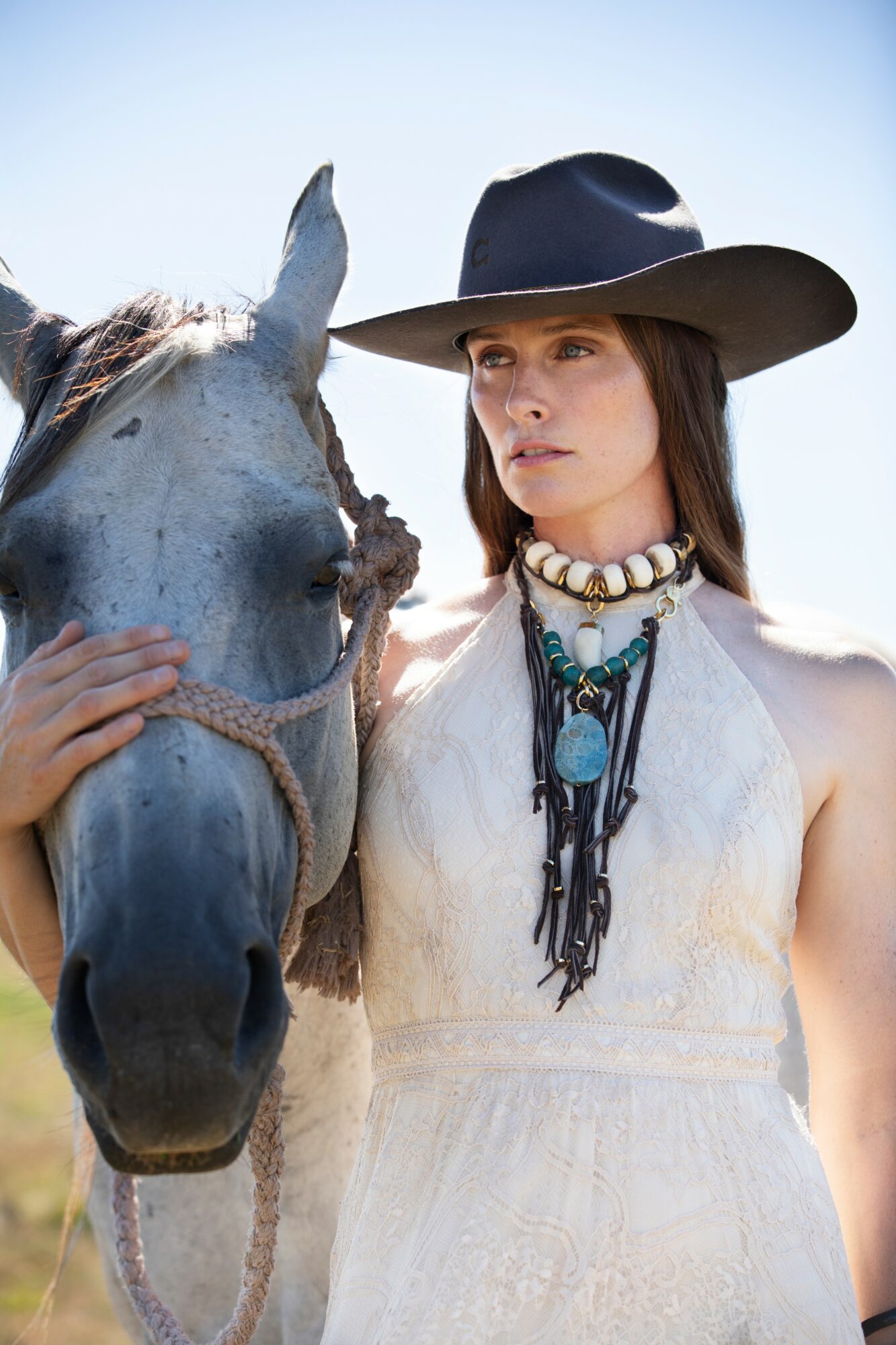 Woman wearing a wide-brimmed hat and layered necklaces stands next to a gray horse, holding its bridle, outdoors on a sunny day.