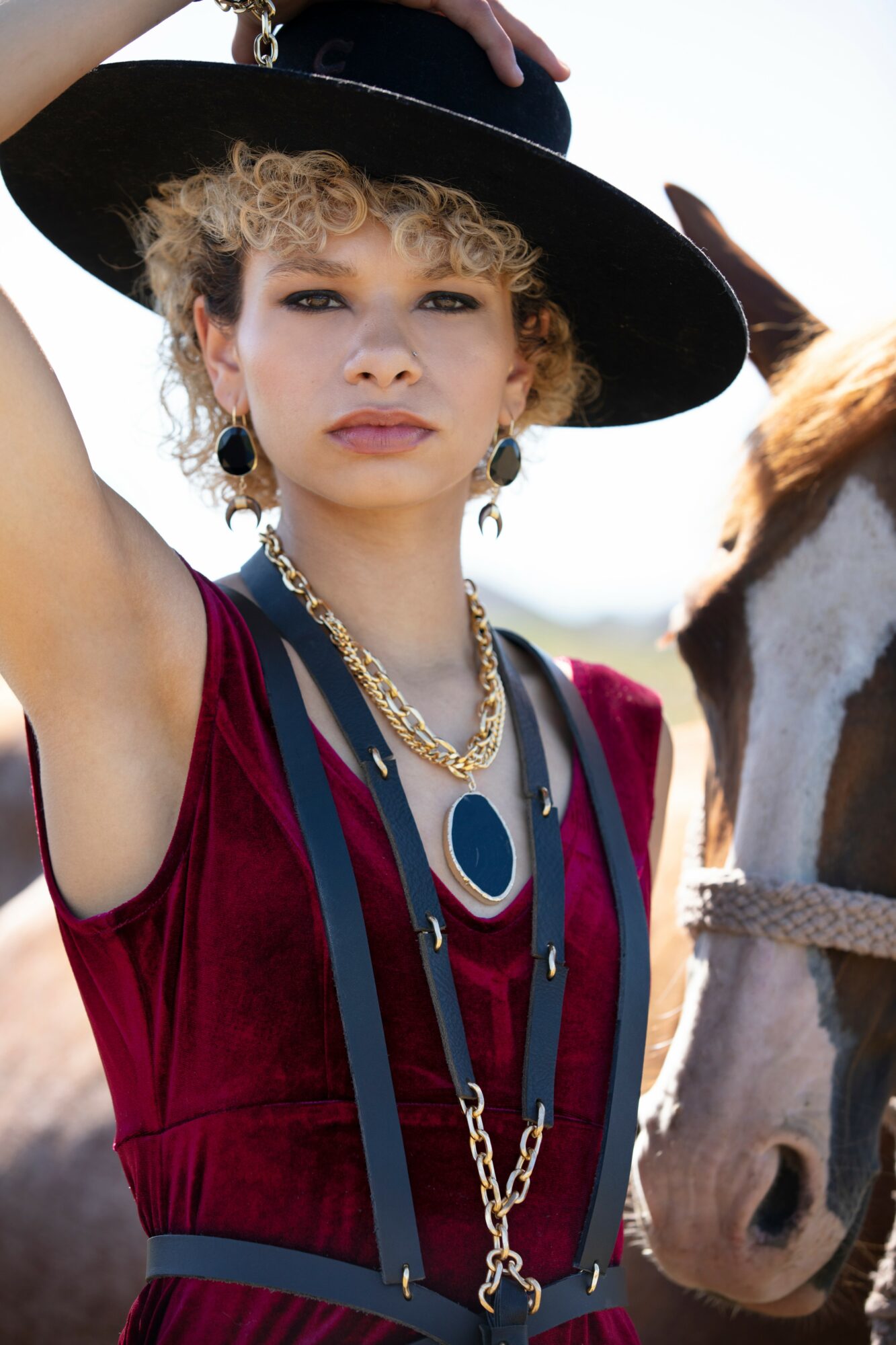 Young woman with curly blonde hair wearing a large black hat, jewelry, and a red and blue outfit, standing outdoors.