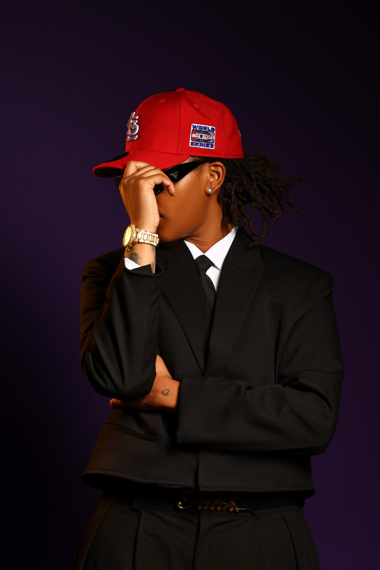 Person wearing a black suit and red cap, covering part of their face, against a dark background.