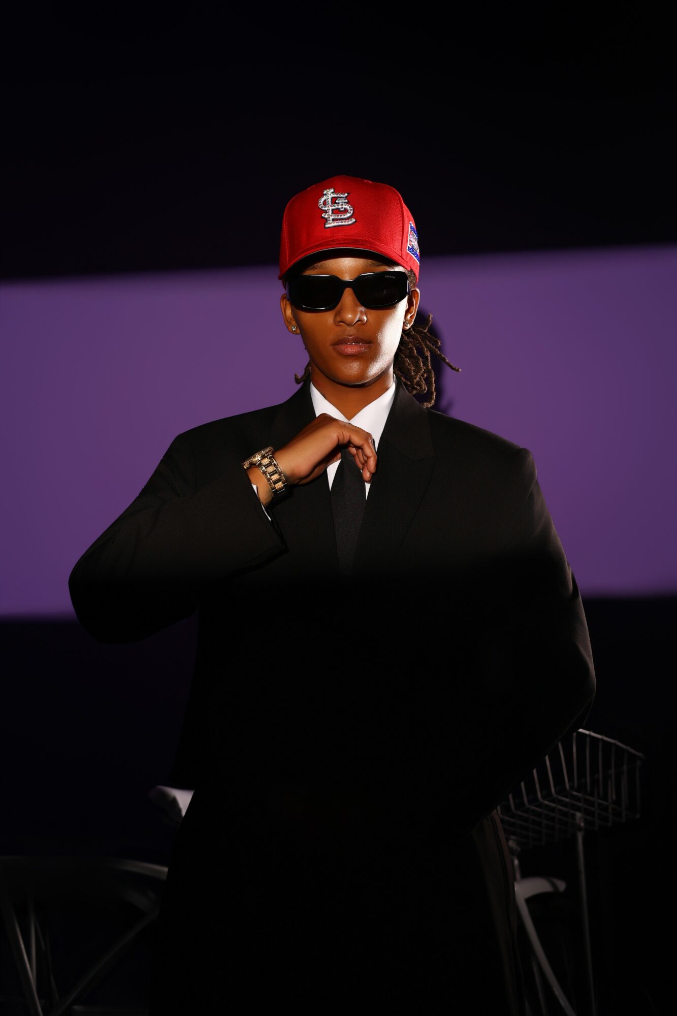 Person wearing sunglasses, red cap, black suit, and jewelry, standing with hand near collar against a dark background.