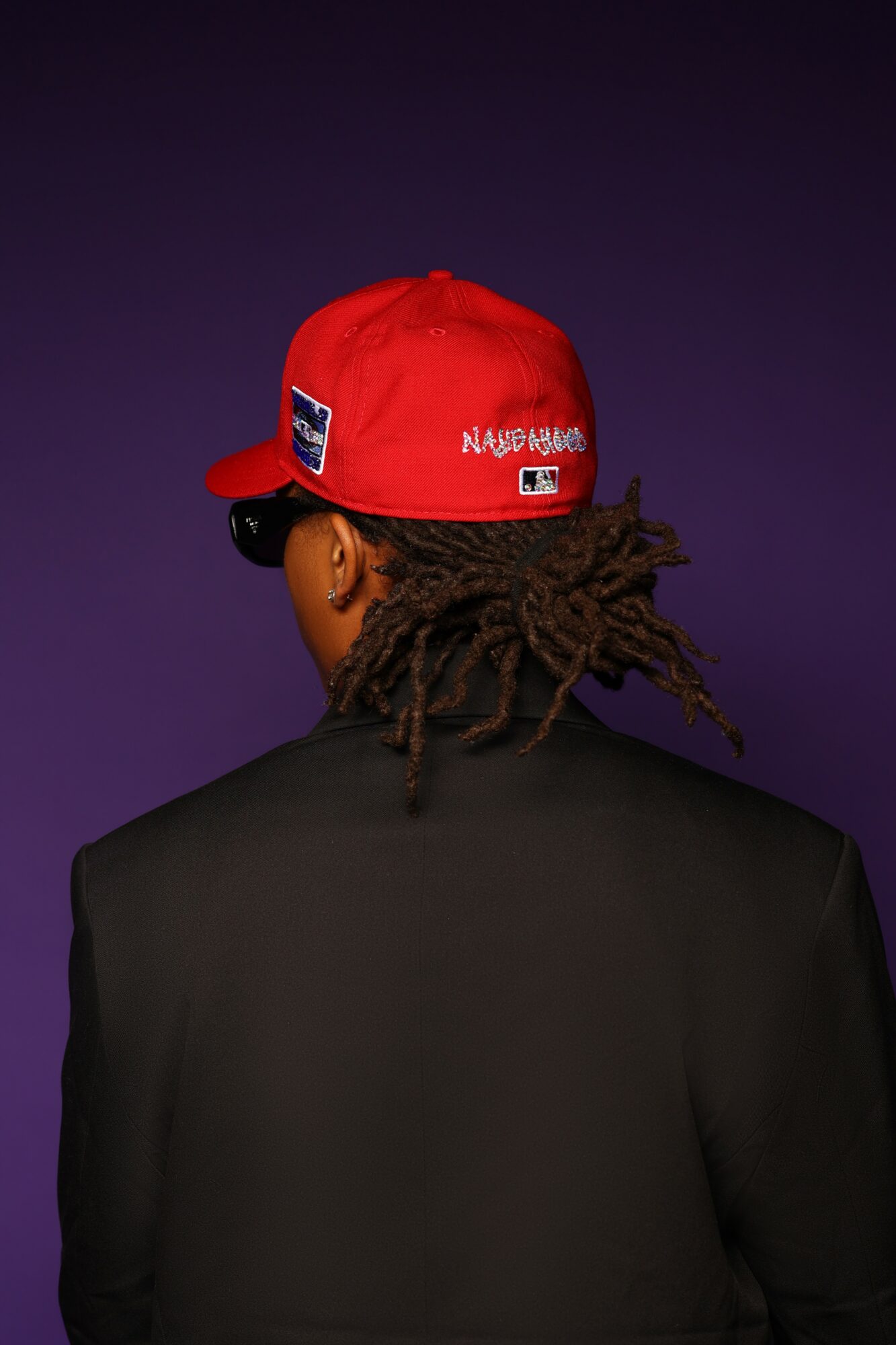 Person with dreadlocks wearing a red cap and black jacket, facing away, against a dark background.