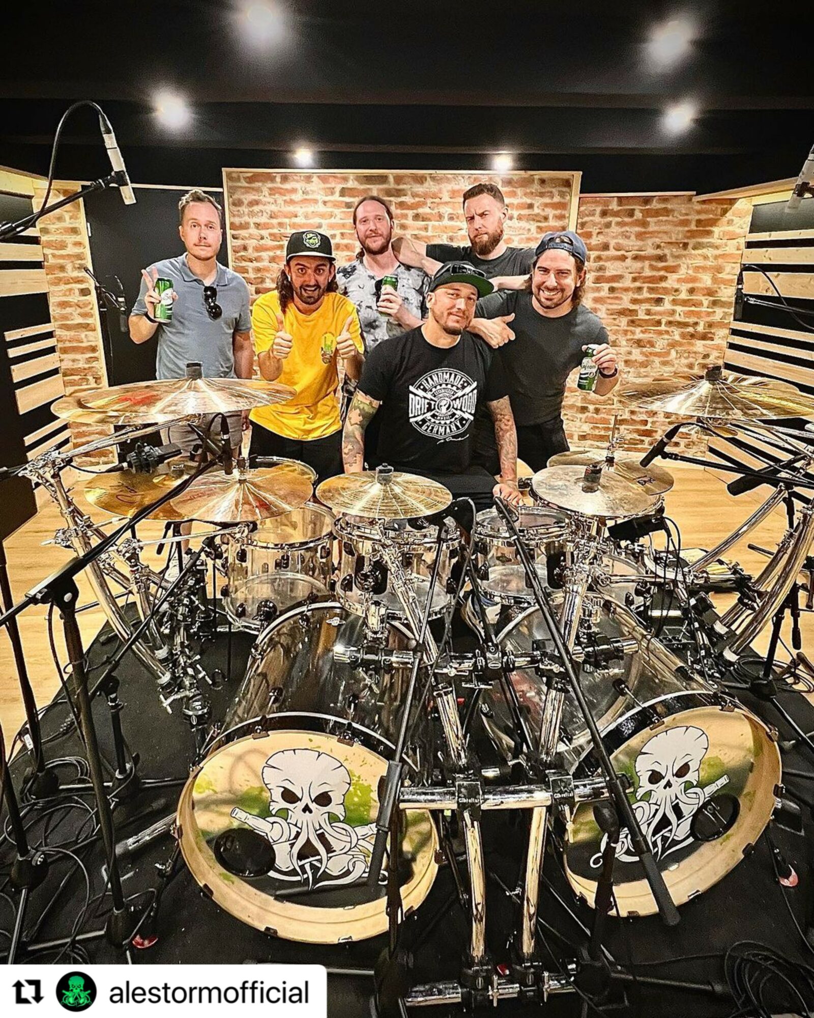 Six people standing behind a drum set in a music studio with brick walls, smiling and posing for the photo.