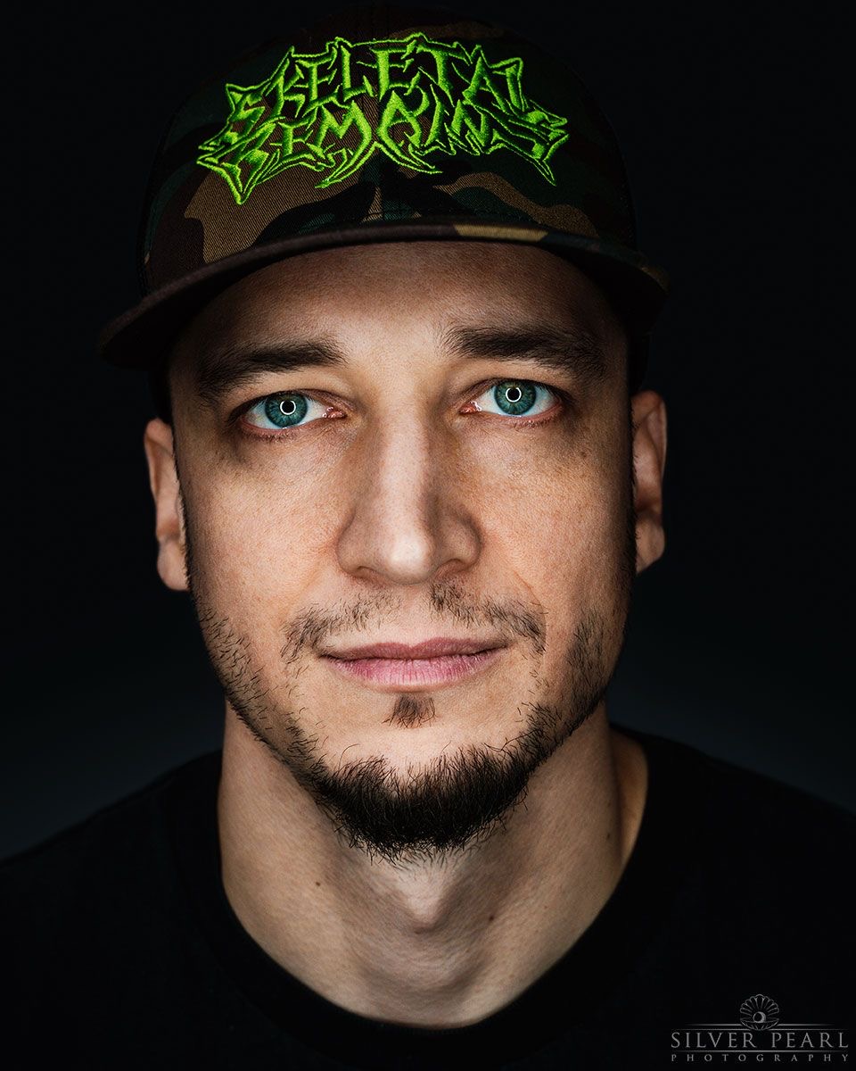 Close-up of a man wearing a cap with green text, blue eyes, and a slight smile, against a dark background.