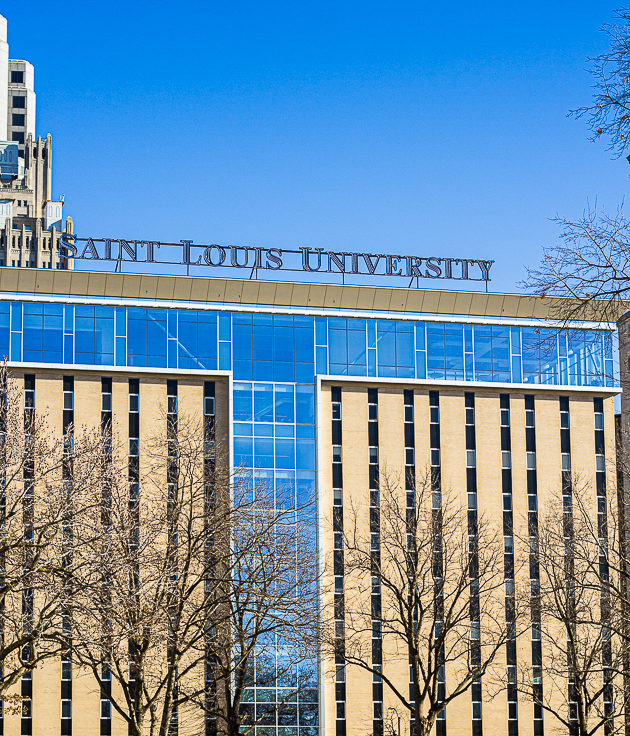Facade of Saint Louis University building with blue glass windows and beige vertical panels, trees in foreground, clear blue sky.