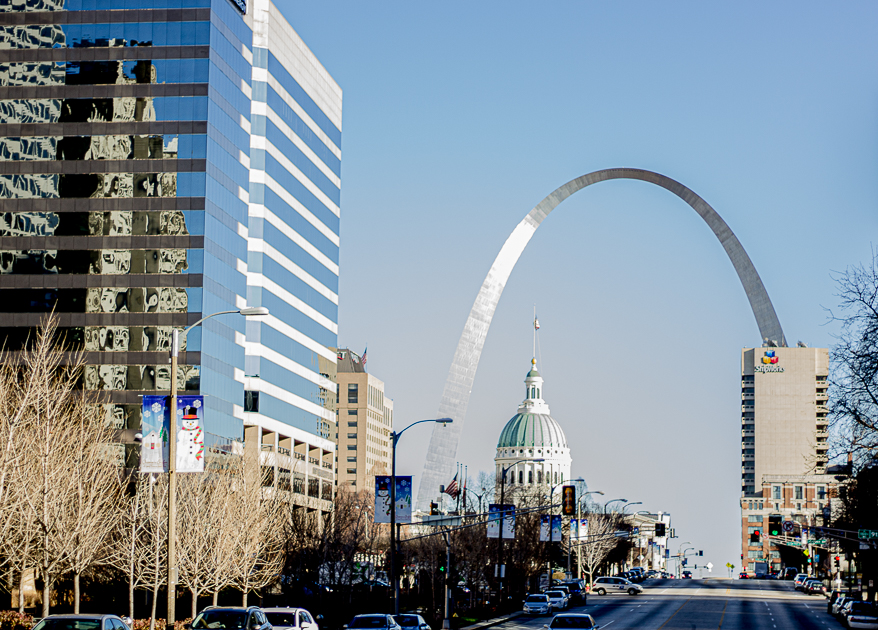 Cityscape with tall buildings, the Gateway Arch, and the Missouri State Capitol in the distance, under a clear blue sky.