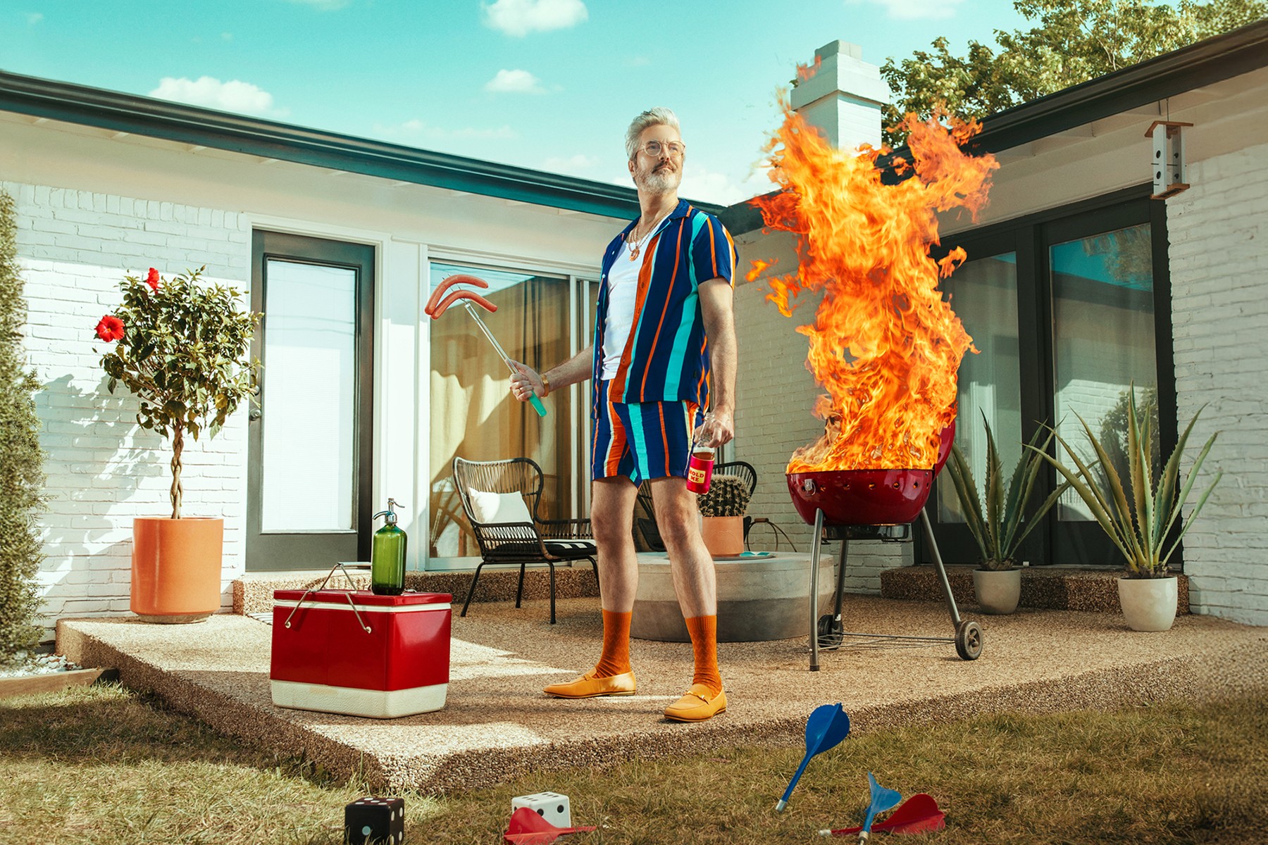 Man standing outdoors next to a barbecue grill with flames, wearing striped shorts and orange shoes, with a cooler and plants nearby.