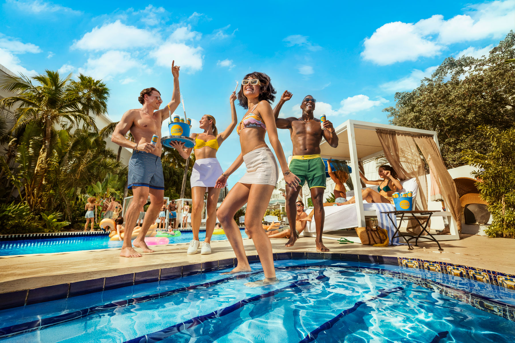 Group of people dancing and playing by a pool outdoors on a sunny day, with trees and blue sky in background.