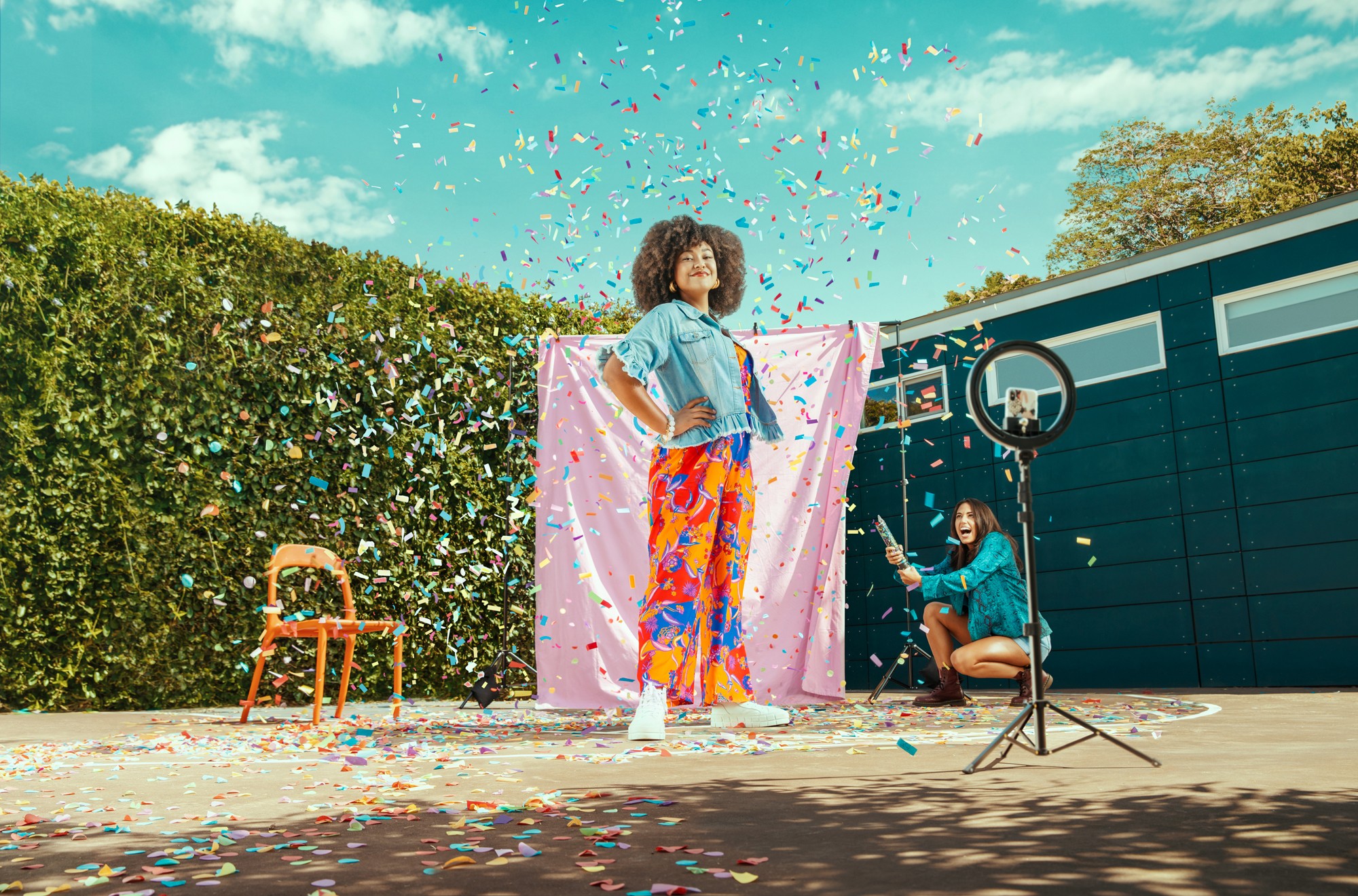 Person standing outdoors with colorful confetti, pink cloth backdrop, and a blue building, surrounded by a hedge and trees.