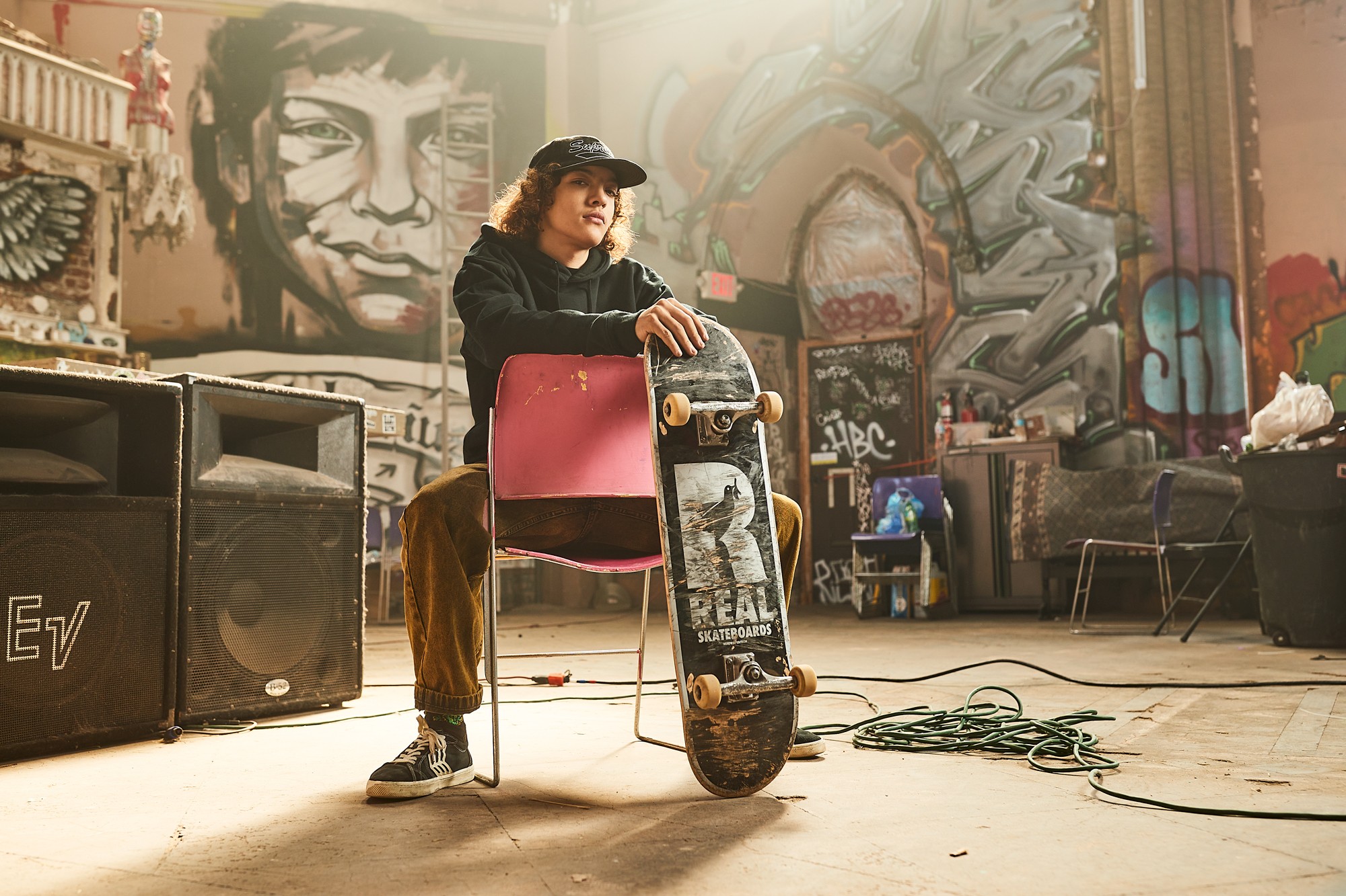Person sitting on a pink chair with a skateboard in an indoor space with graffiti-covered walls, equipment, and chairs.