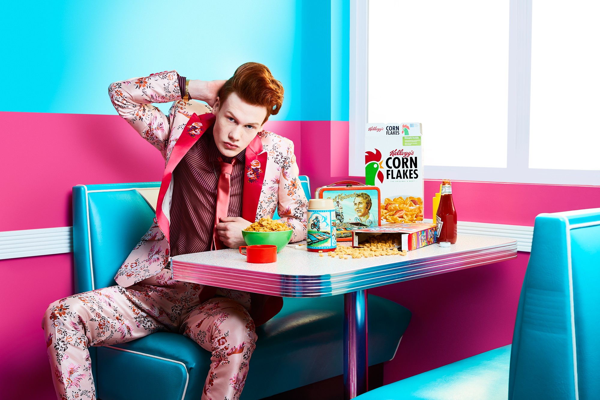 Person sitting in a colorful room with cereal boxes on the table, wearing a patterned suit, with one hand on head.