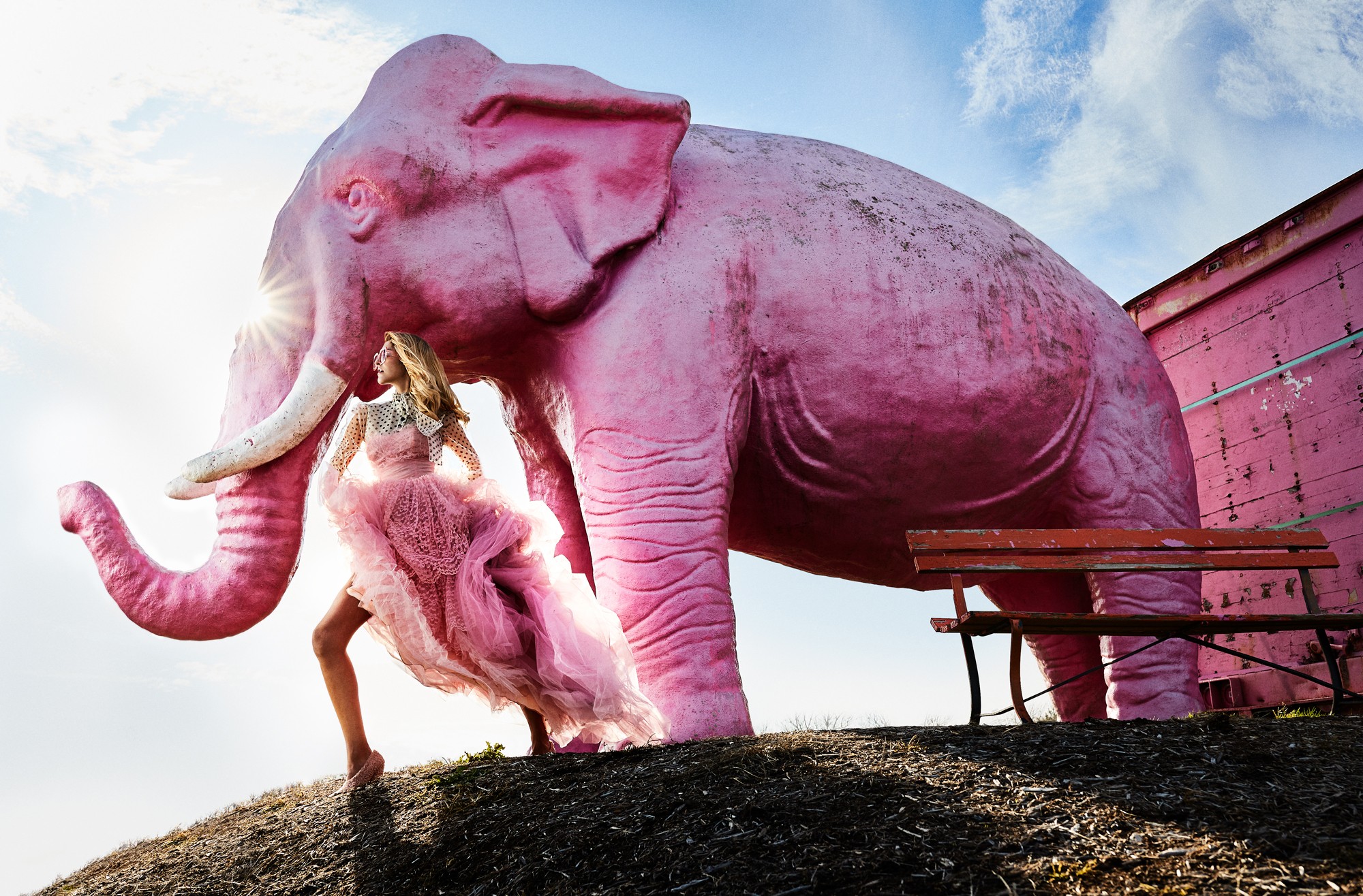 A person in a pink dress stands next to a large pink elephant sculpture outdoors, with a blue sky and clouds in the background.