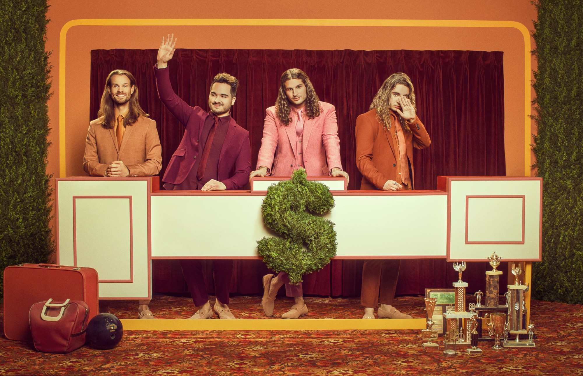 Four people stand behind a game show podium with a large green plant in front, trophies, and a red bag nearby.