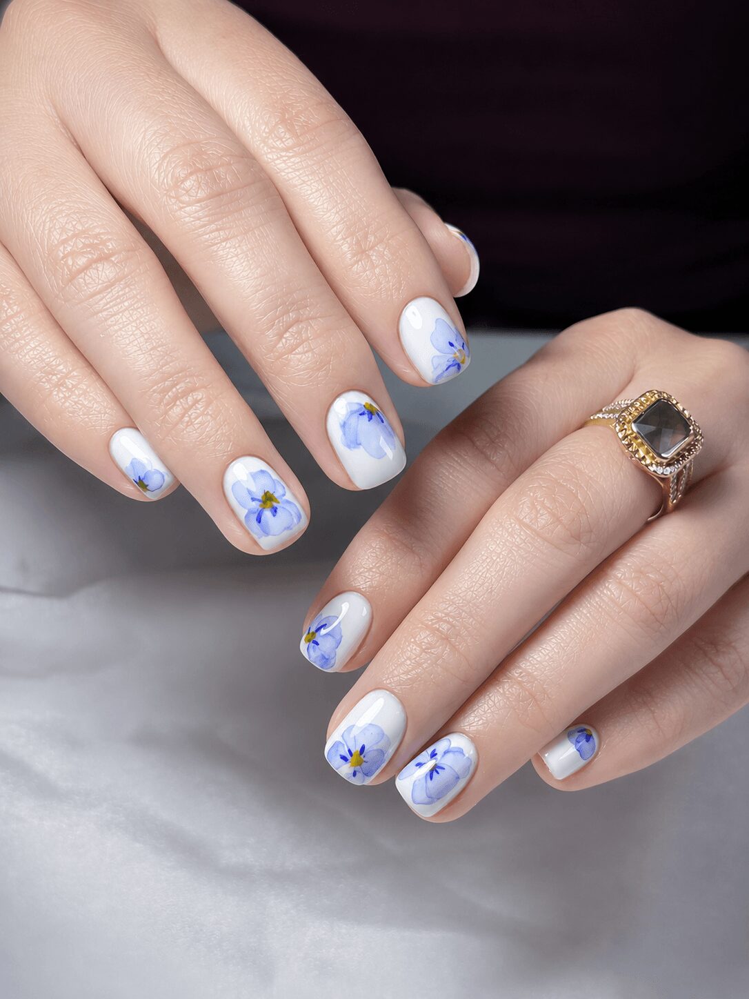 Hands with painted nails featuring mountain and sky designs, one hand wearing a gold ring with a dark stone, neutral background.