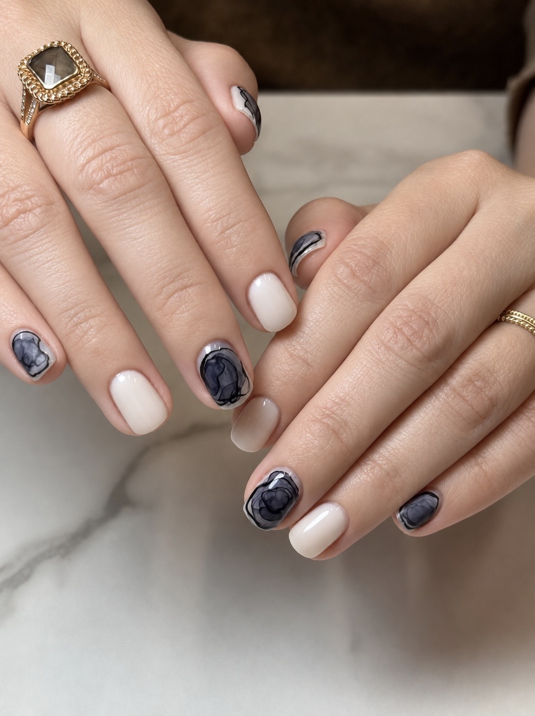 Two hands with manicured nails and rings, one hand with a gold ring and the other with a silver ring, resting on a marble surface.