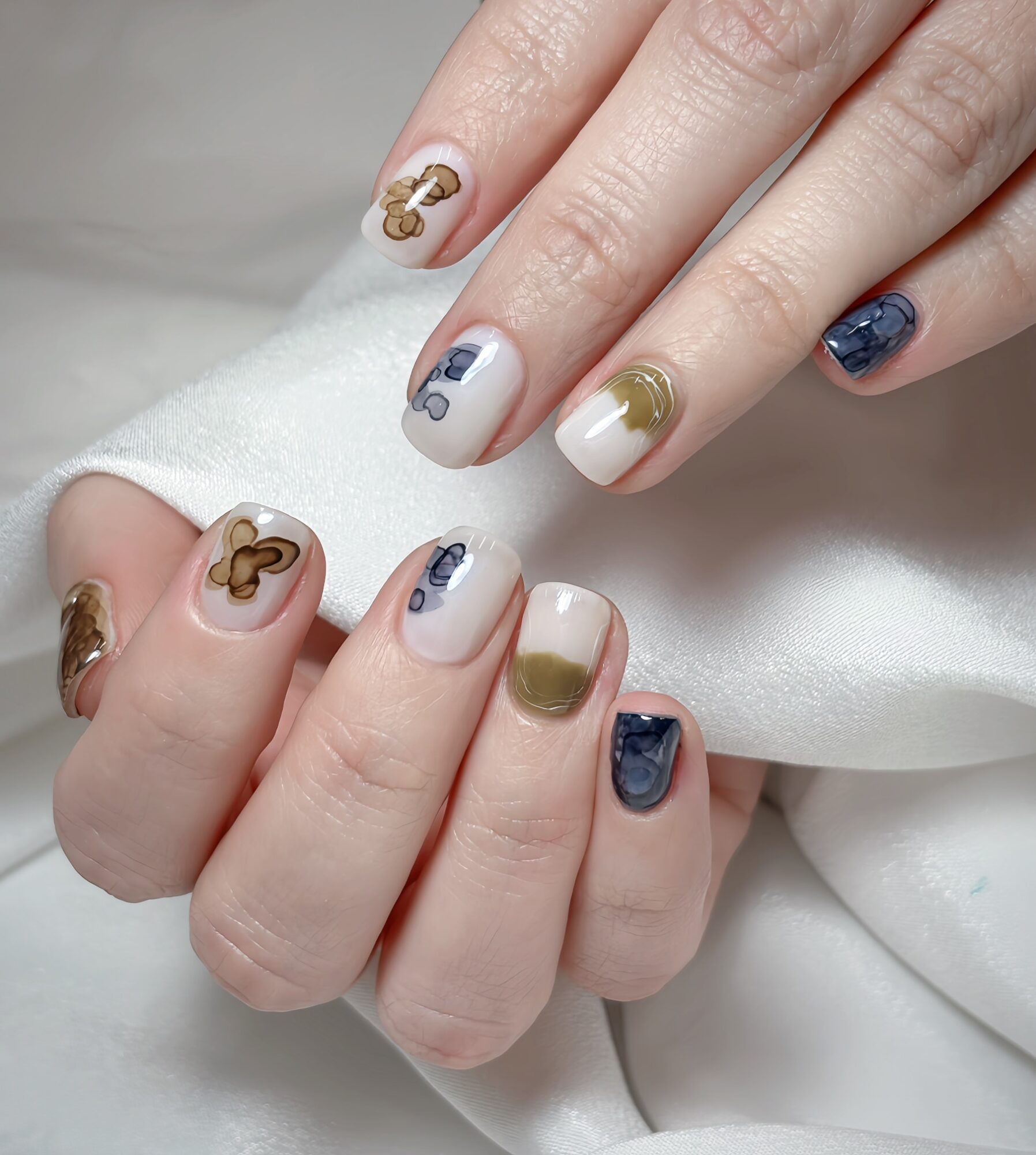 Hands with decorated nails, featuring gold, blue, and beige designs, resting on white fabric.