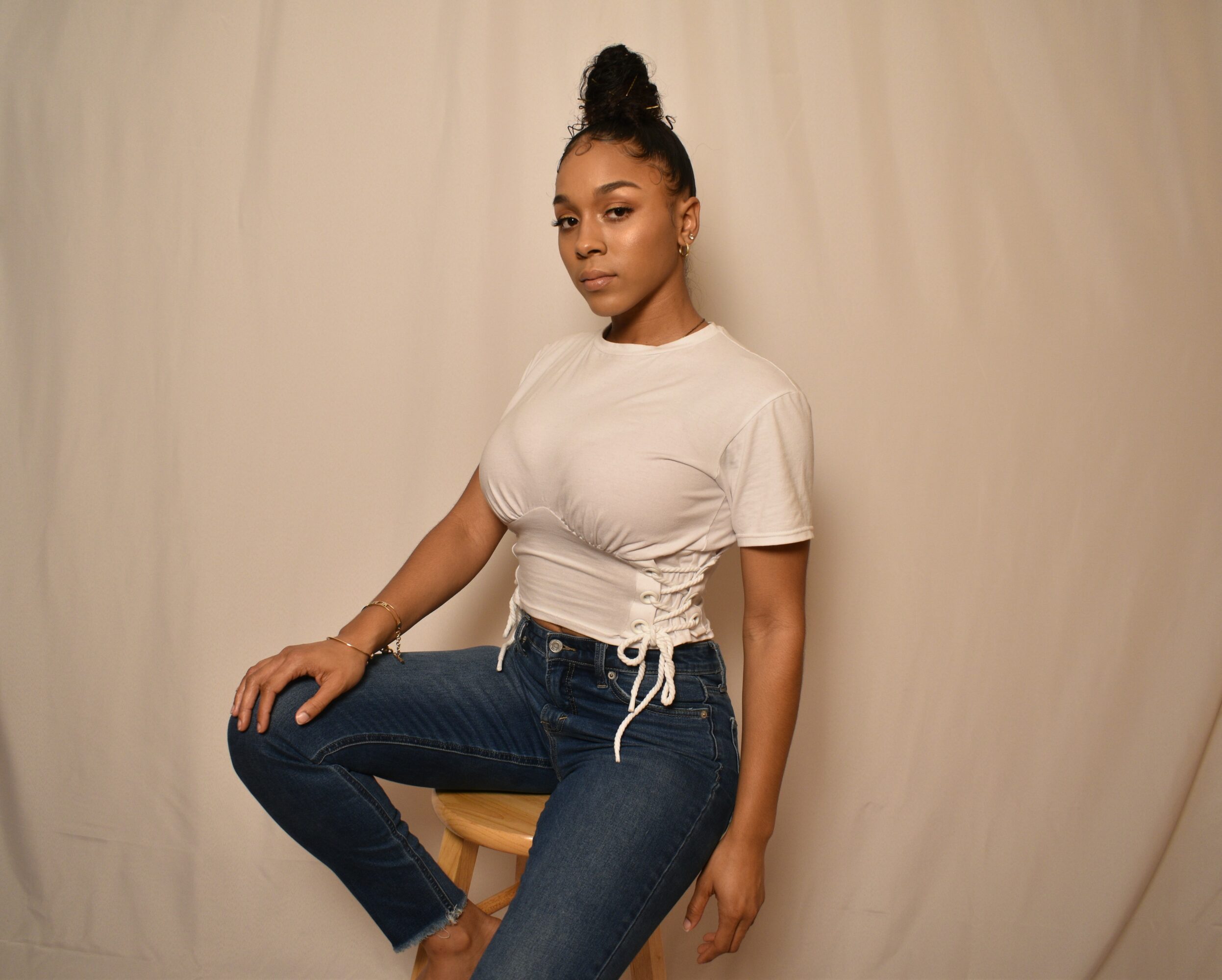 Young woman sitting on a stool, wearing a white t-shirt and jeans, against a plain background.