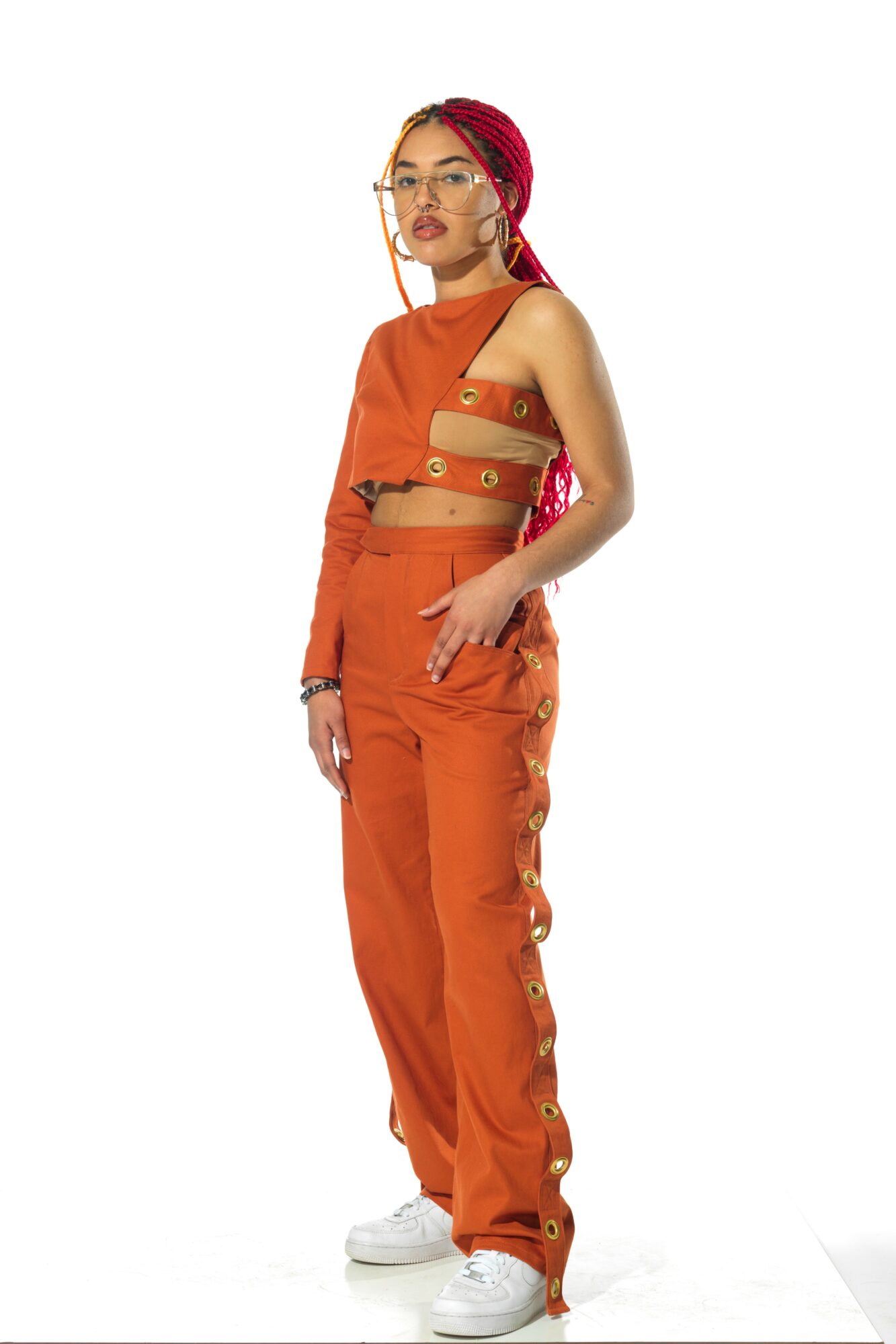 Young woman with pink hair and glasses wearing an orange outfit, standing with one hand in pocket, facing forward.