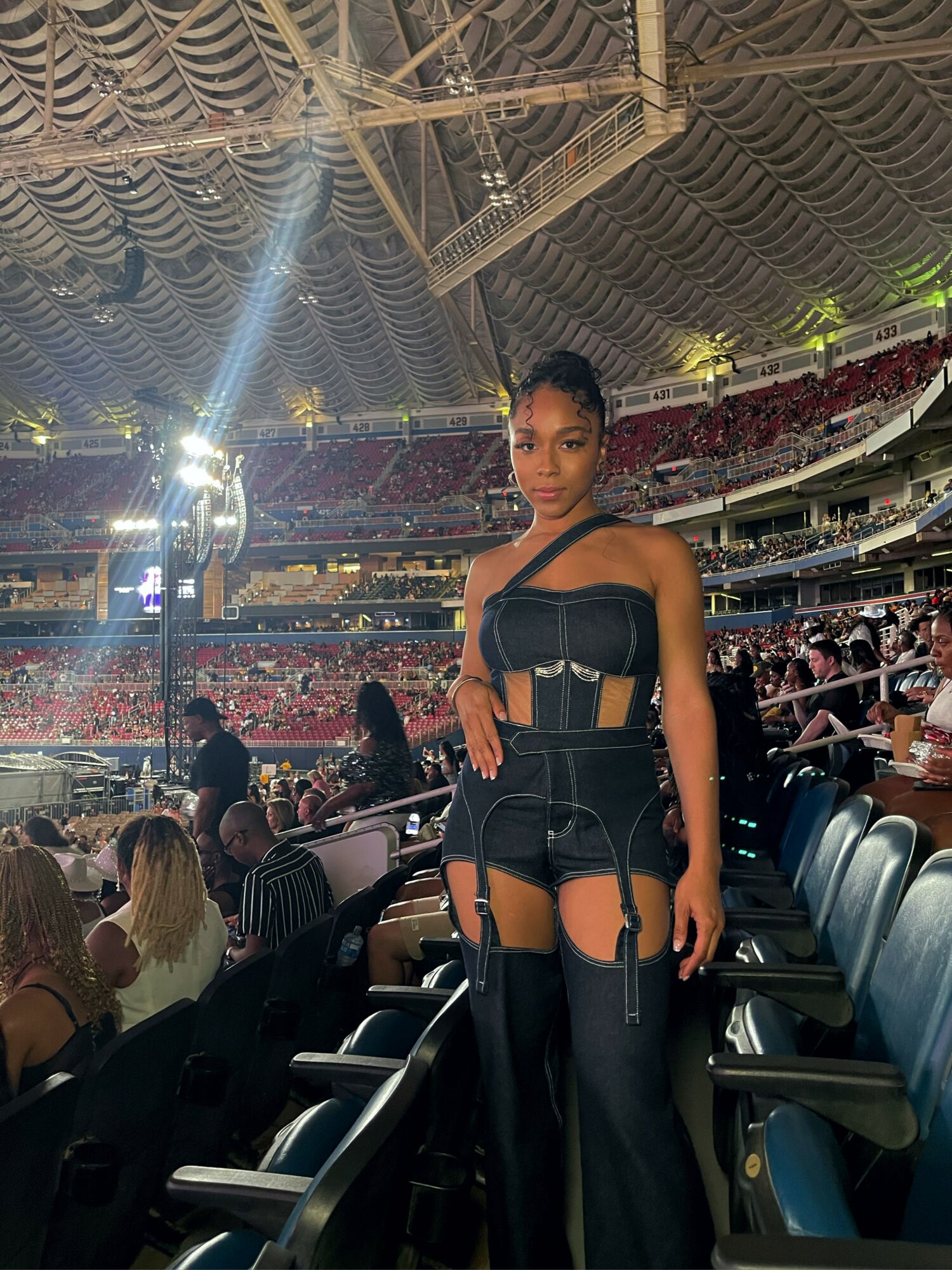 Young woman in black outfit standing in stadium with audience and bright lights, ceiling with draped fabric overhead.