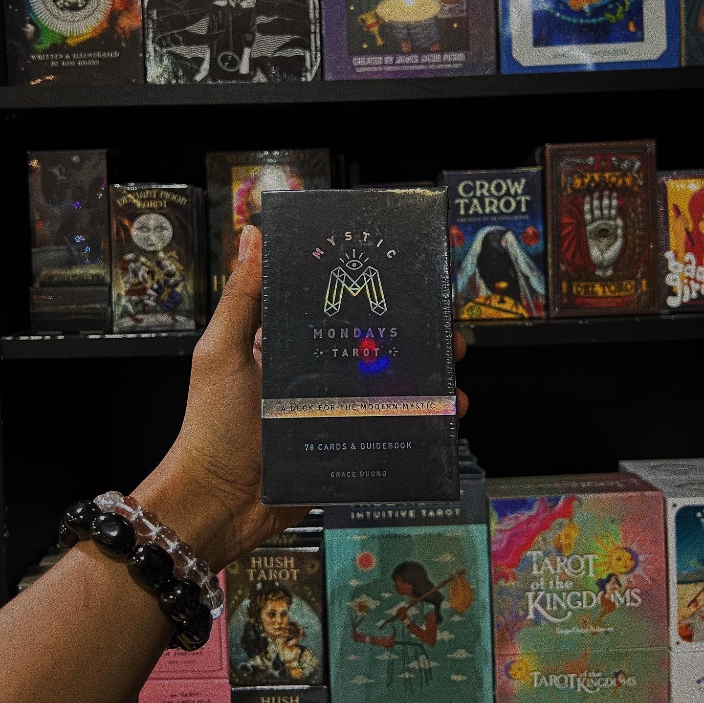 Person holding a tarot card deck in a store, with shelves of tarot and oracle card decks behind.