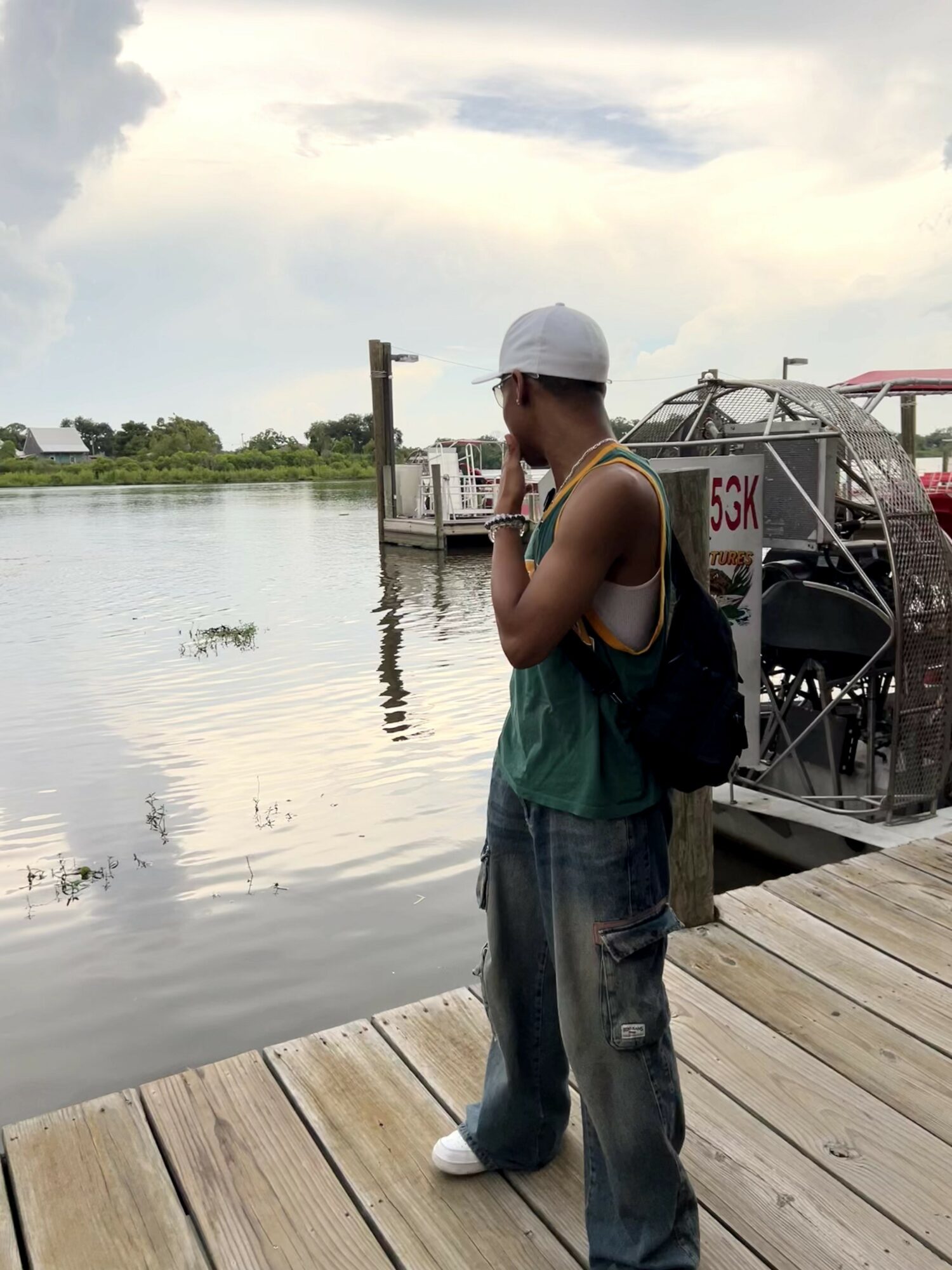 Person standing on a wooden dock near water, wearing a white cap, sunglasses, and casual clothing, with boats in background.