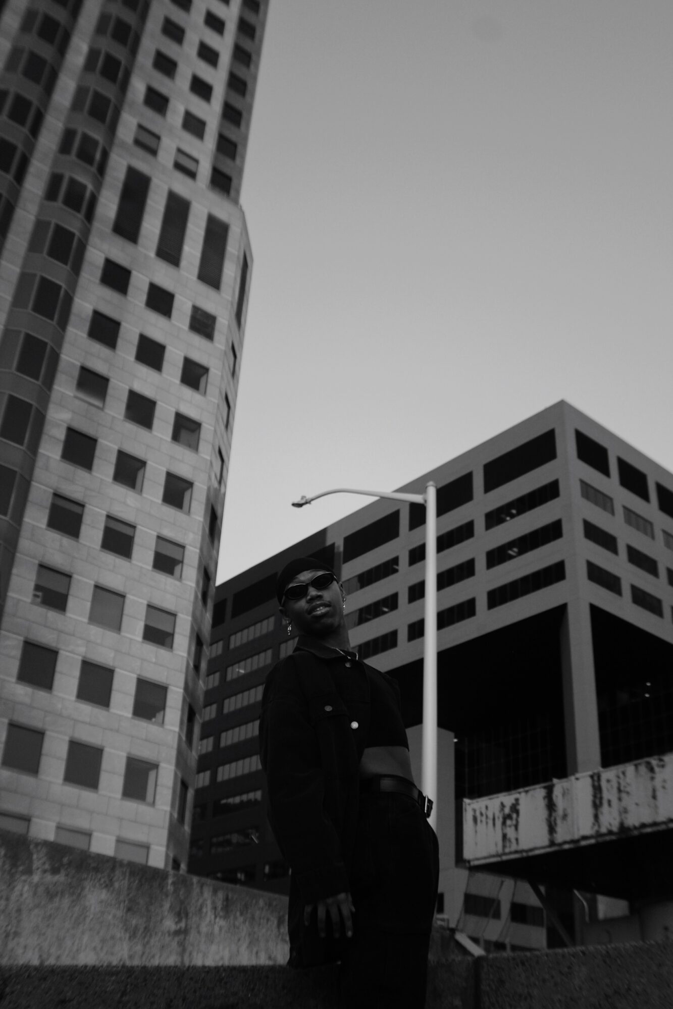 Person standing in front of tall buildings with a streetlamp, black and white, urban scene.