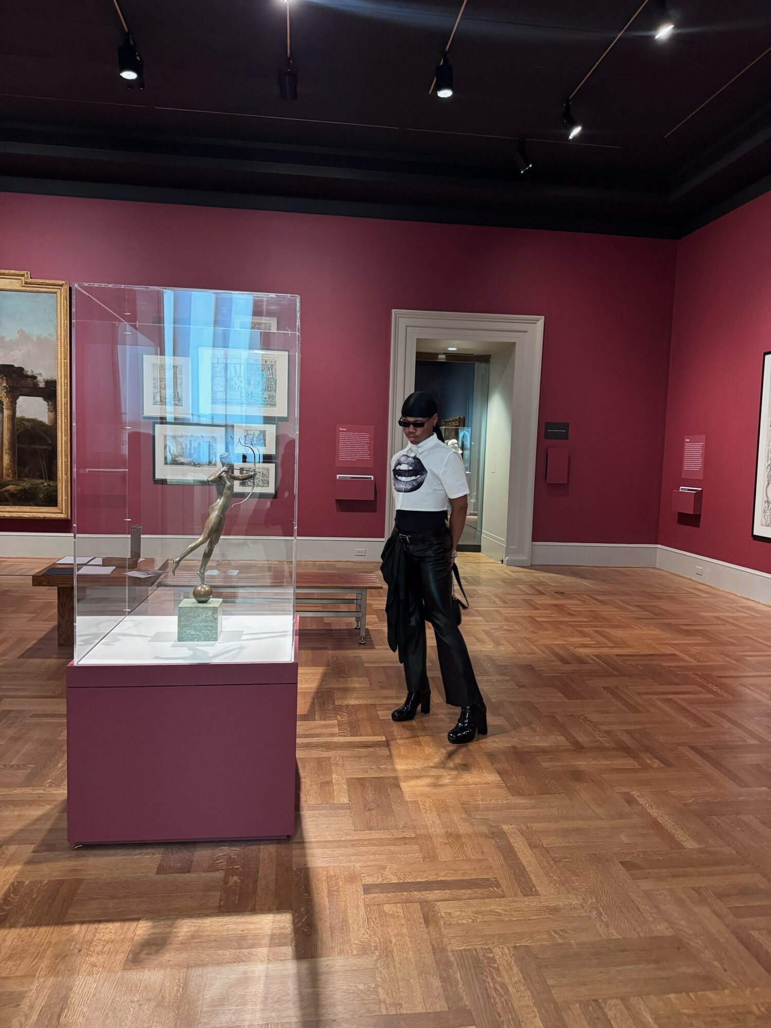 Person standing in an art gallery with pink walls, wooden floor, and display cases, looking at artwork.