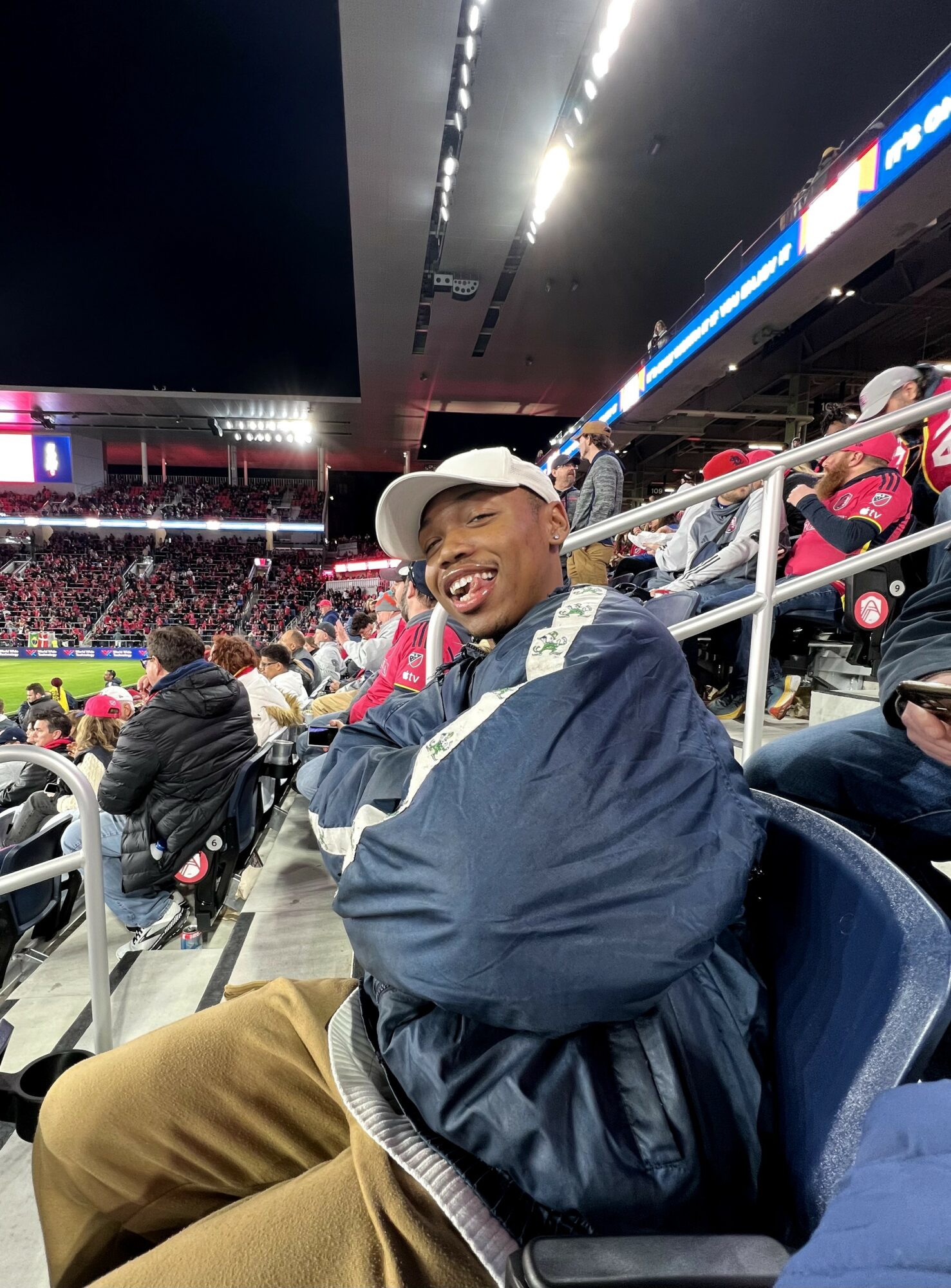 Person smiling, wearing a white cap and dark jacket, sitting in stadium seats at a sports event, with a crowd and field in background.