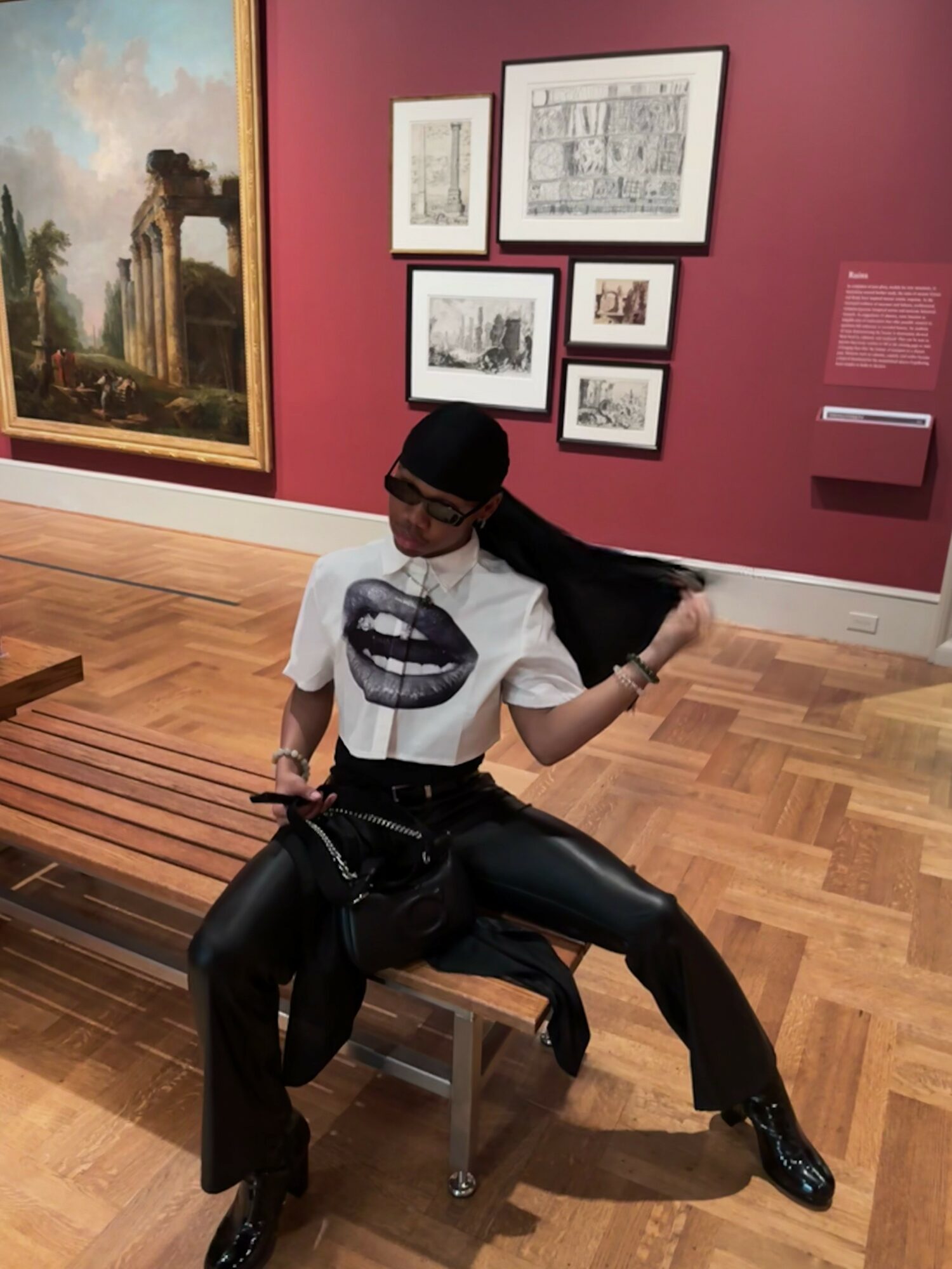 Person sitting on a bench in an art gallery, wearing a white t-shirt with a mouth graphic, black pants, and glasses, holding their hair.