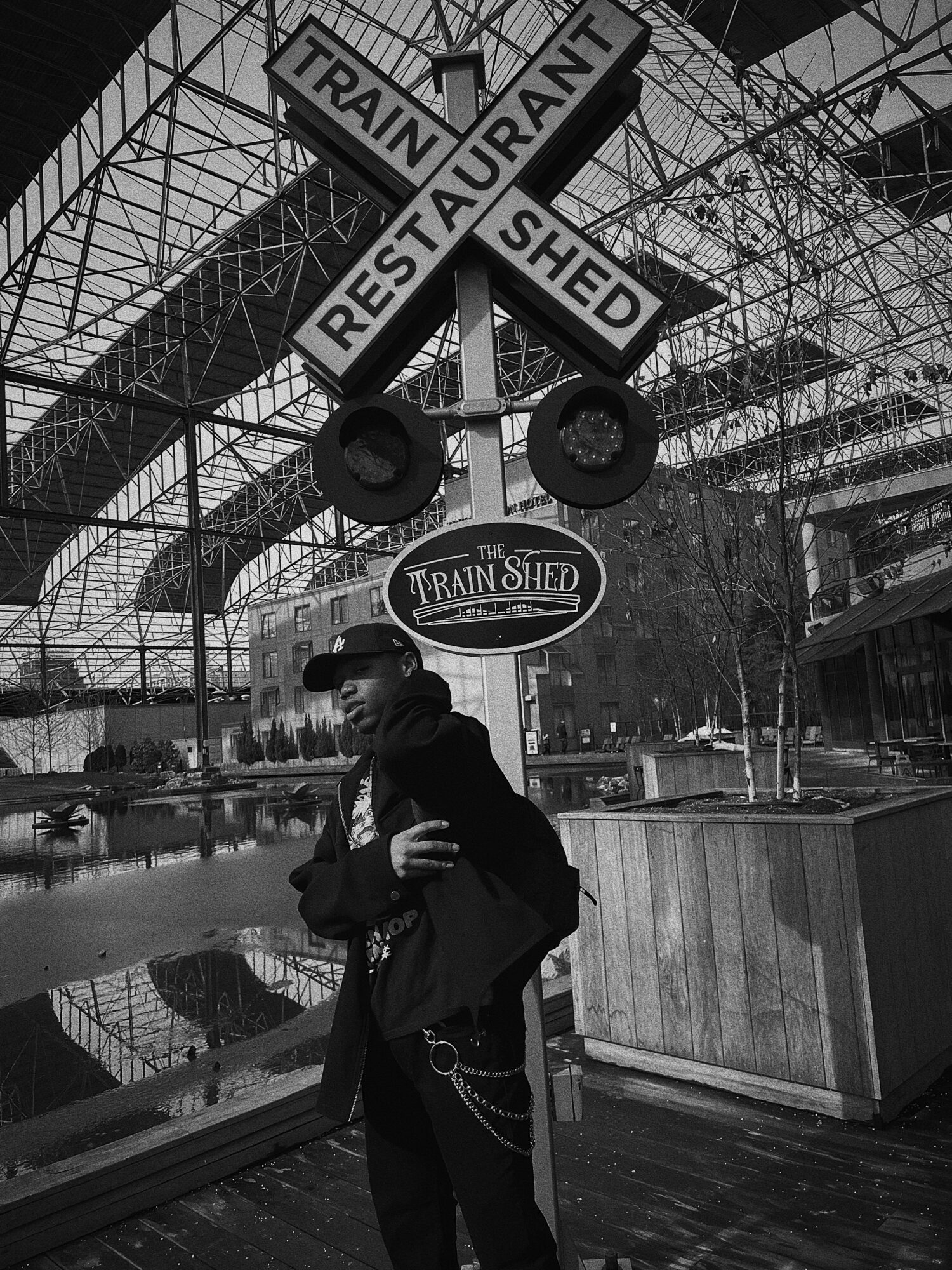 Person standing indoors near a sign for a train shed and restaurant, with glass ceiling and reflections on the floor.