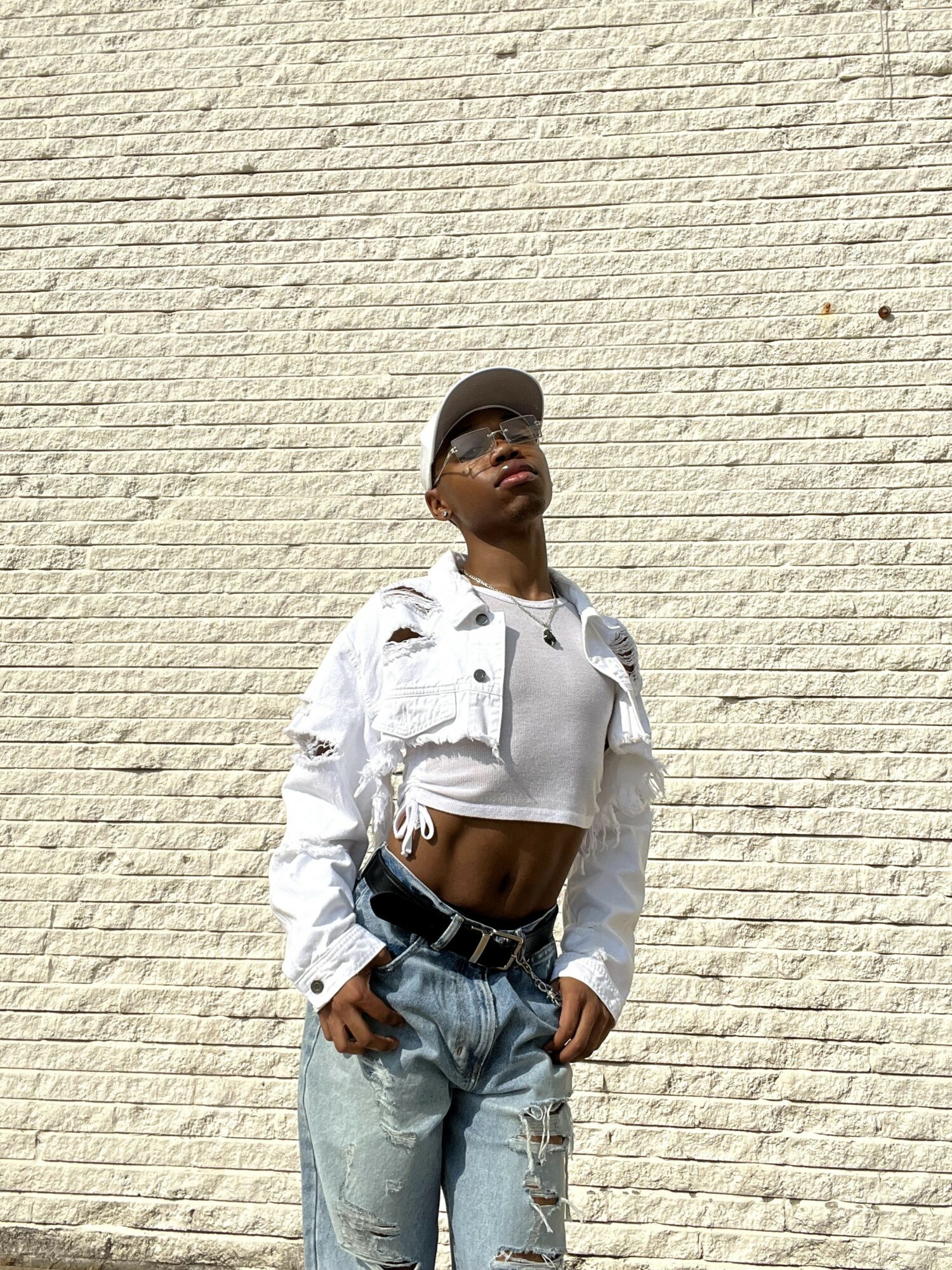 Person wearing a white cropped jacket, light blue jeans, black belt, sunglasses, and a cap, standing against a textured wall.