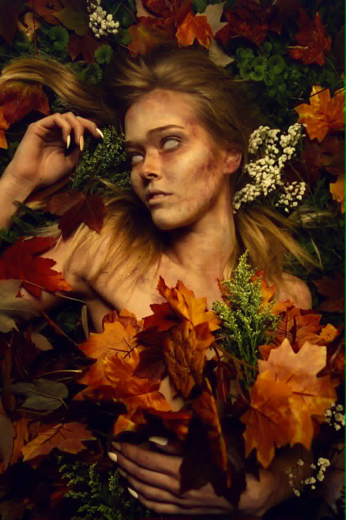 Young woman with long hair lying among colorful autumn leaves and white flowers, gazing to the side.