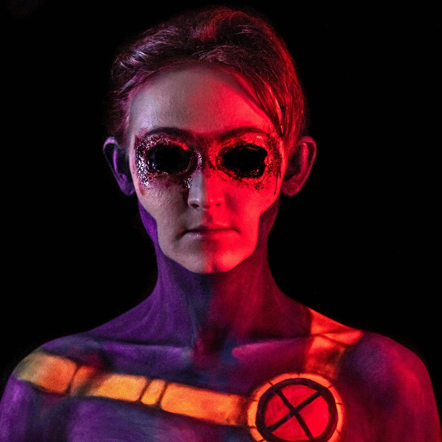 Person with face paint and colorful body paint, dark background, intense expression, short hair, dramatic lighting.