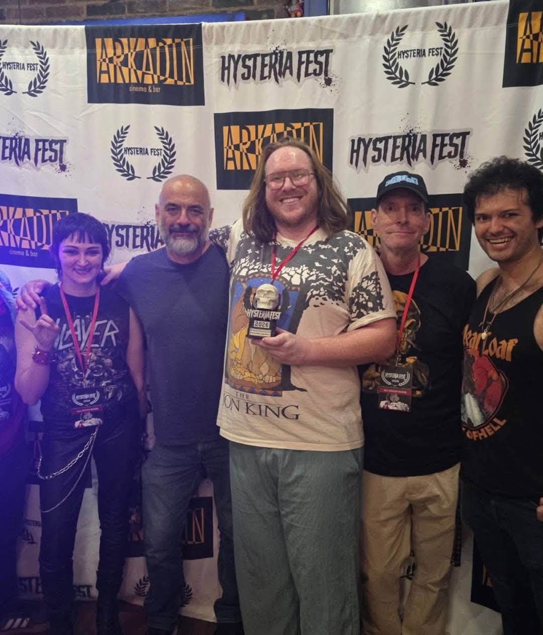 Group of five people smiling in front of a Hysteria Fest banner, some holding medals, indoors.