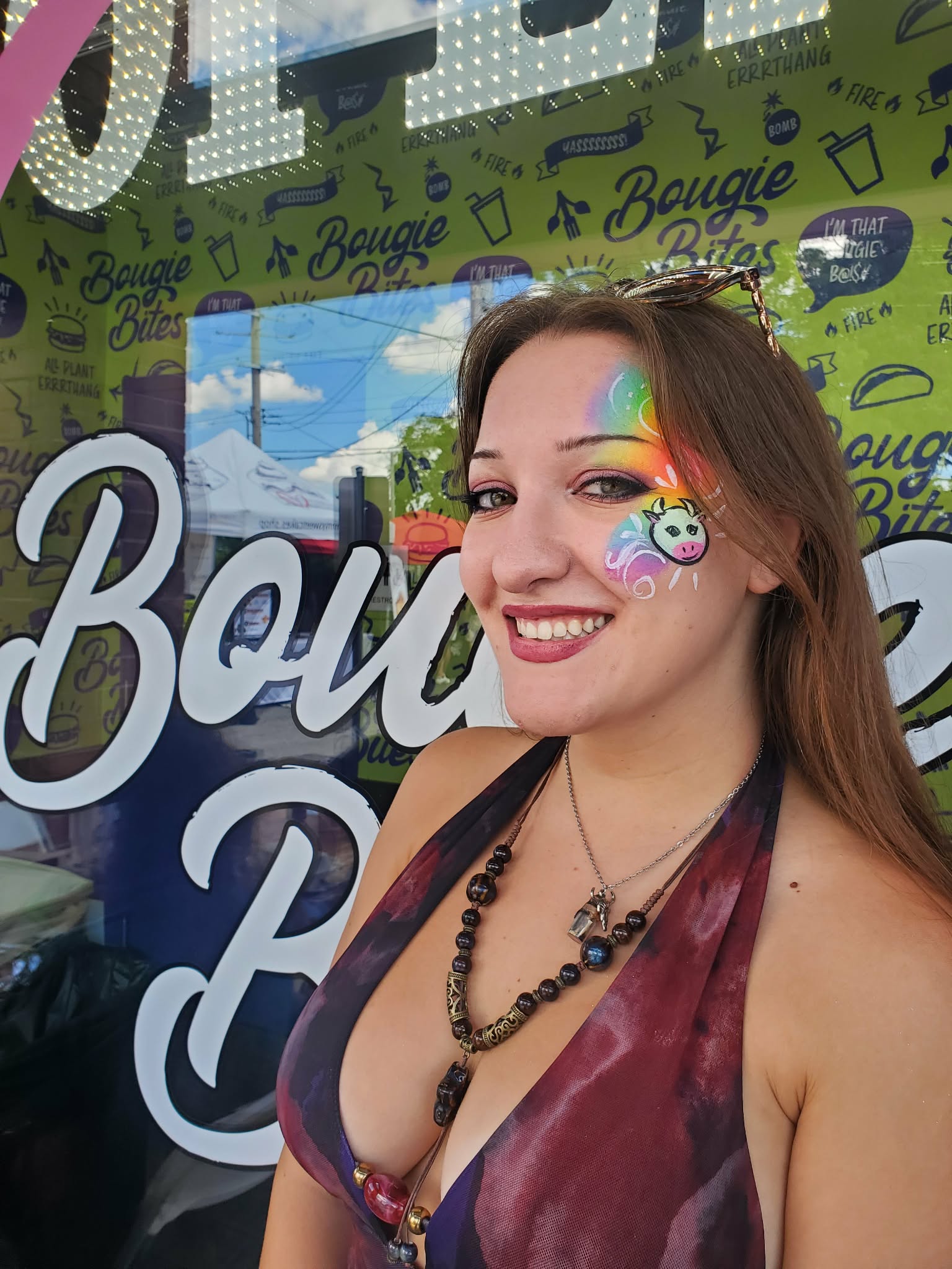 Smiling woman with rainbow face paint and a beaded necklace, standing in front of a colorful backdrop with text and graphics.