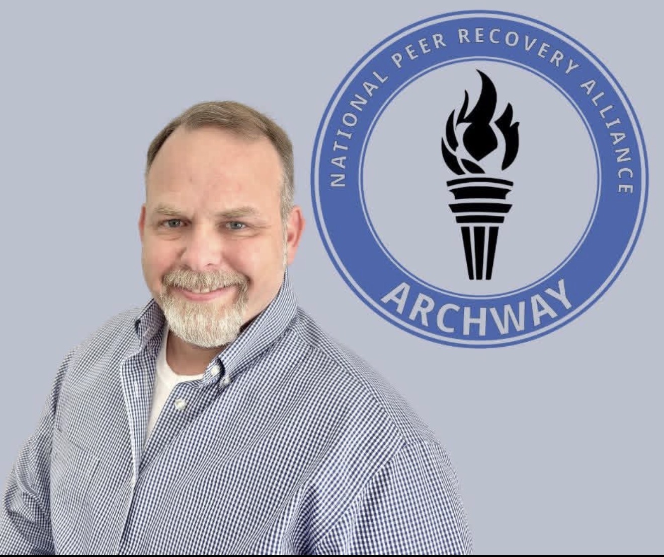 Man with a beard and short hair smiling, standing in front of a logo with a torch and the words 'ARCHWAY' and 'National Peer Recovery Alliance'.