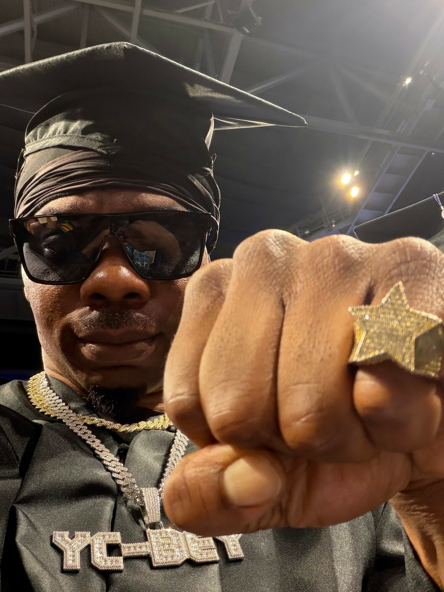 Person wearing sunglasses, graduation cap, and a star ring, showing a fist with a gold star ring, in an indoor setting.