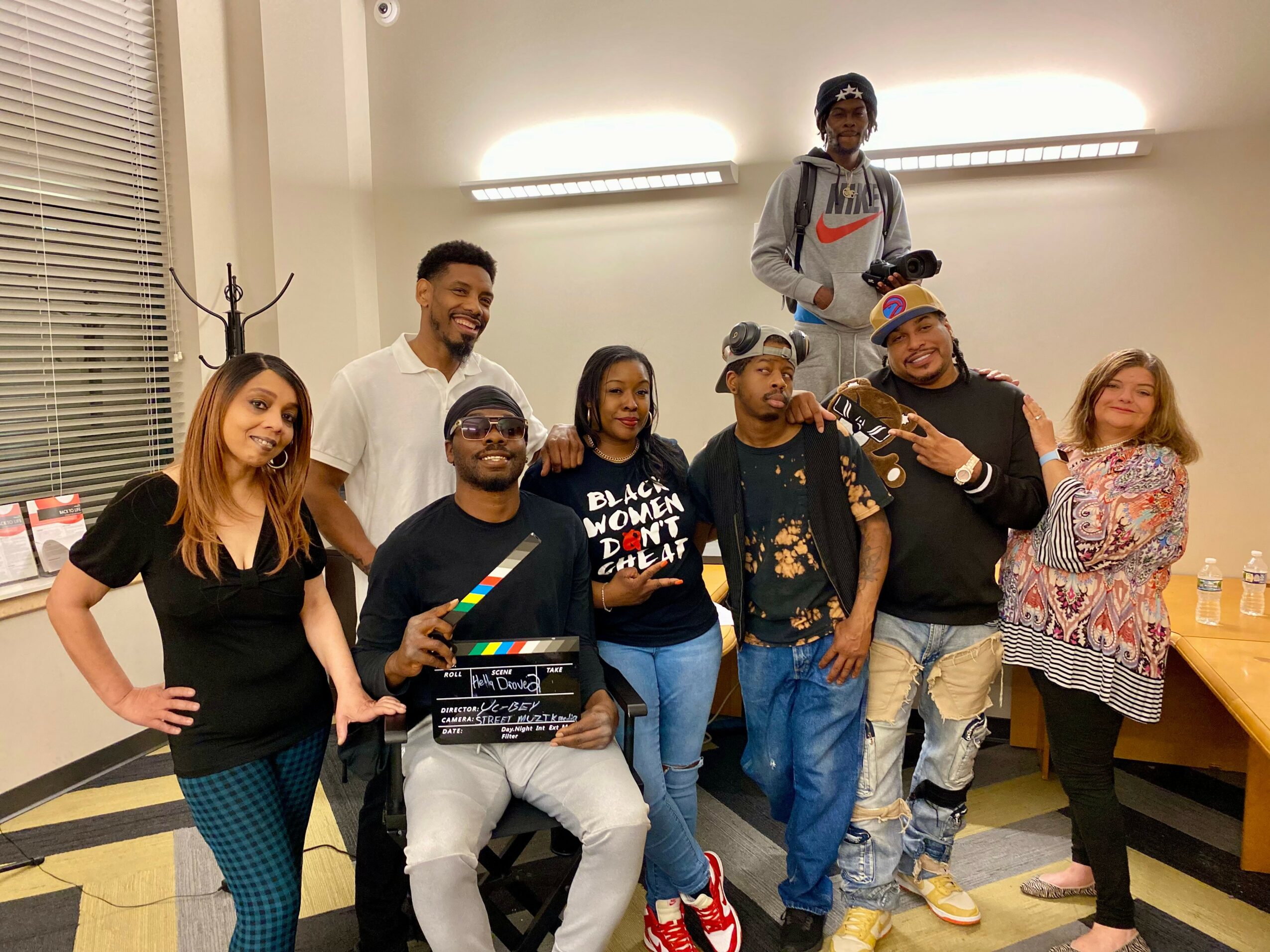 Group of eight people posing indoors, some holding drinks and a clapperboard, smiling and standing in a room with bright lighting.