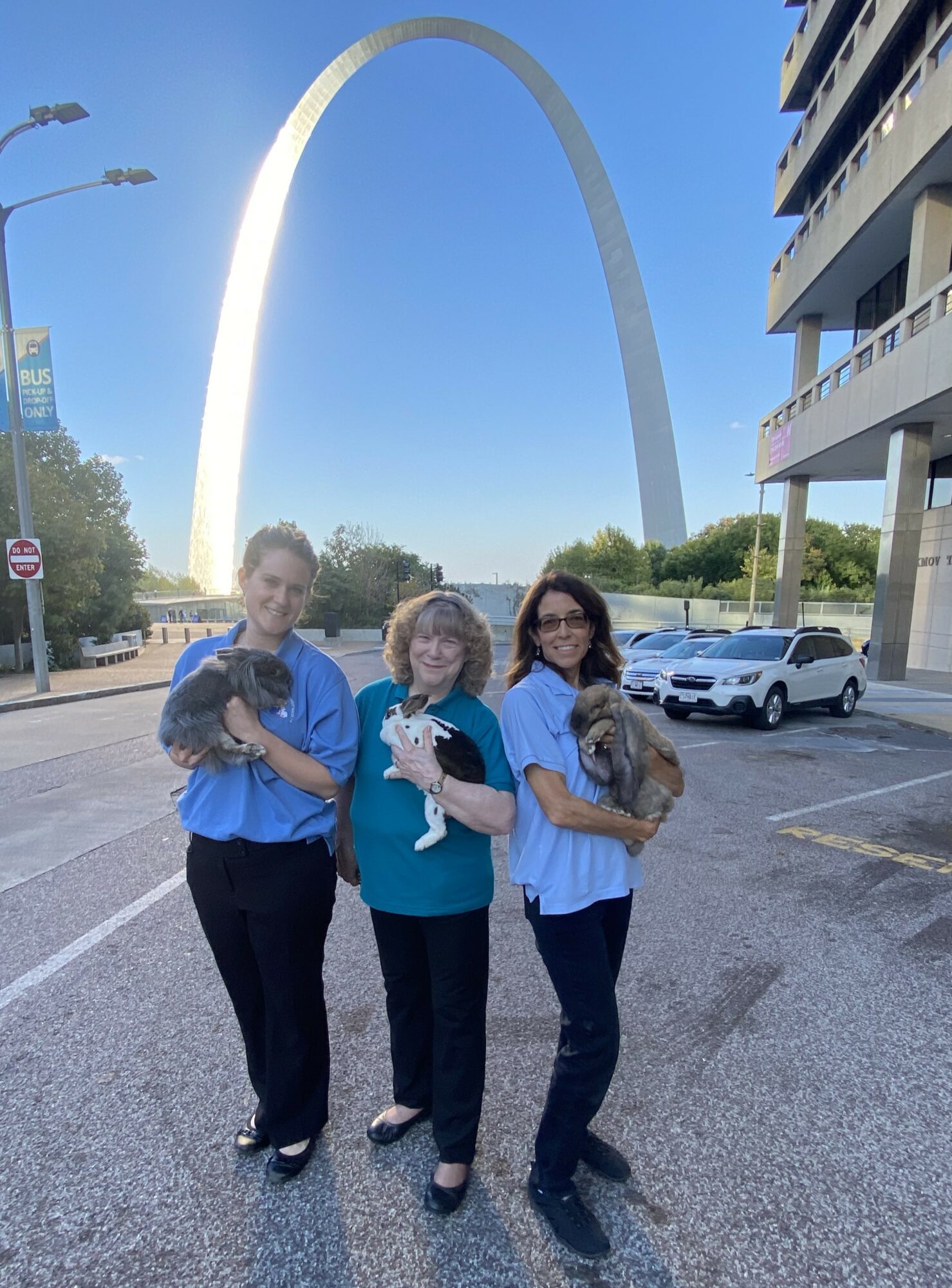Three women standing outdoors holding cats, with the Gateway Arch in the background on a sunny day.