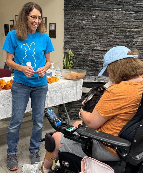 Woman smiling and talking to a person in a wheelchair, indoors with a table and plants in the background.