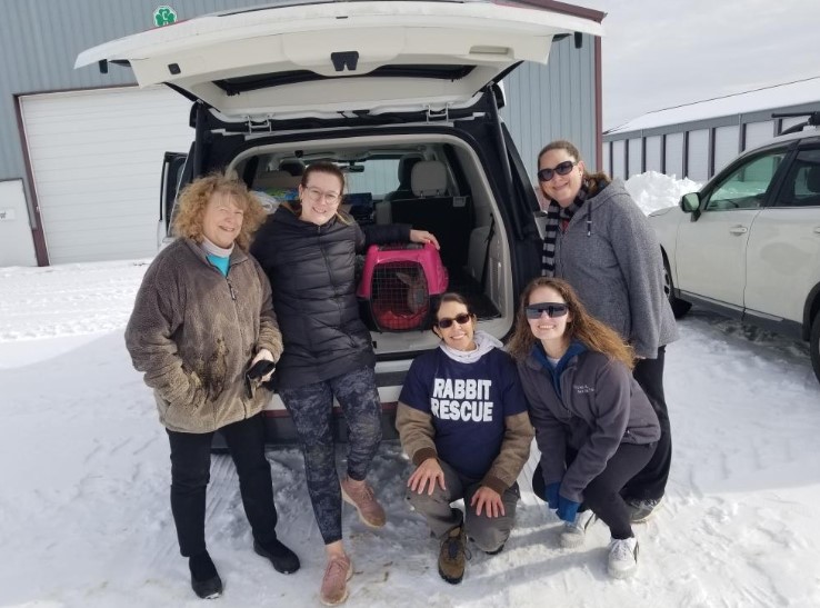 Five people standing in front of an open vehicle trunk outdoors in snow, some wearing sunglasses and winter clothing.