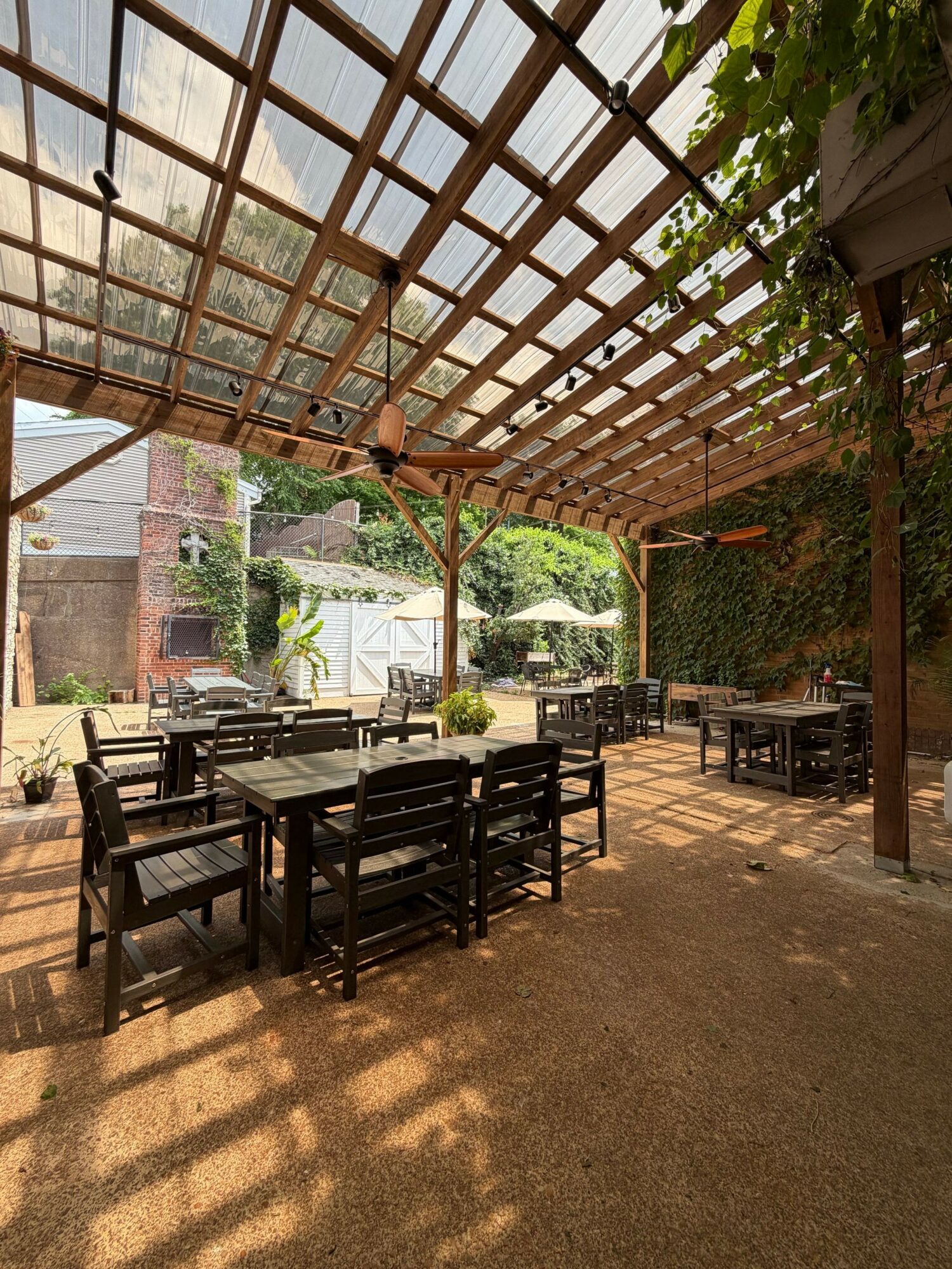 Outdoor seating area with black chairs and tables under a wooden pergola, with trees and buildings in the background.
