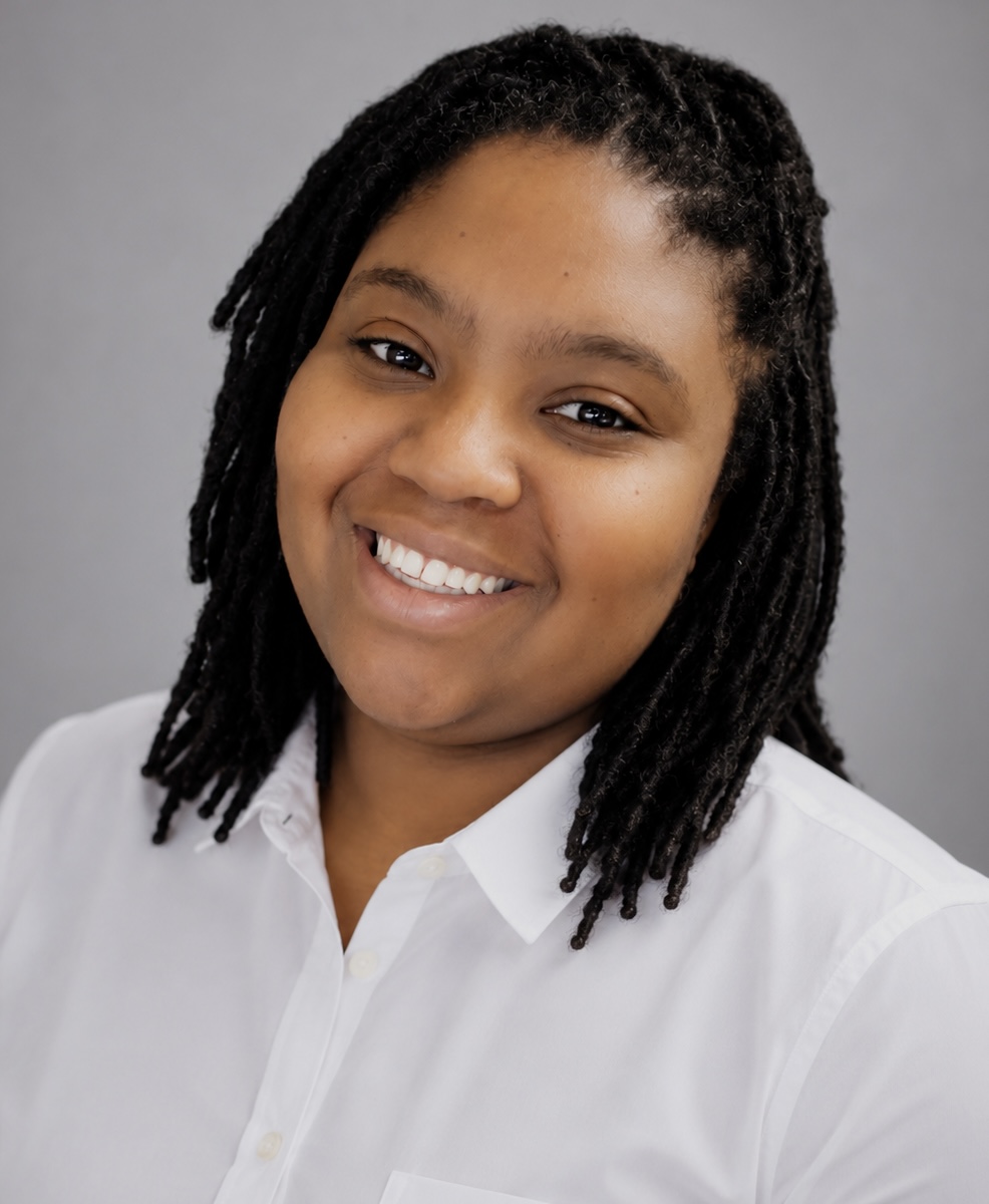 Portrait of a smiling woman with shoulder-length dreadlocks wearing a white shirt against a gray background.