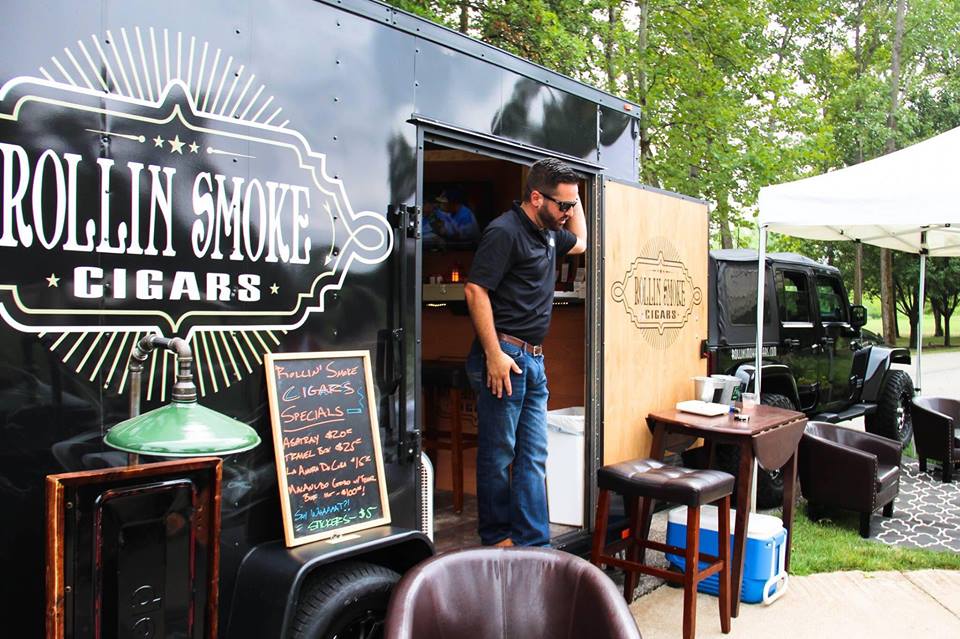 Man standing outside a cigar truck with a chalkboard menu, surrounded by outdoor seating and greenery.