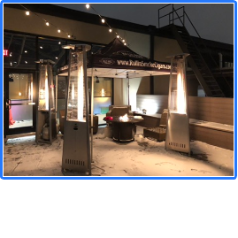 Outdoor patio with heaters, string lights, and seating area at night, snow on the ground, building in background.