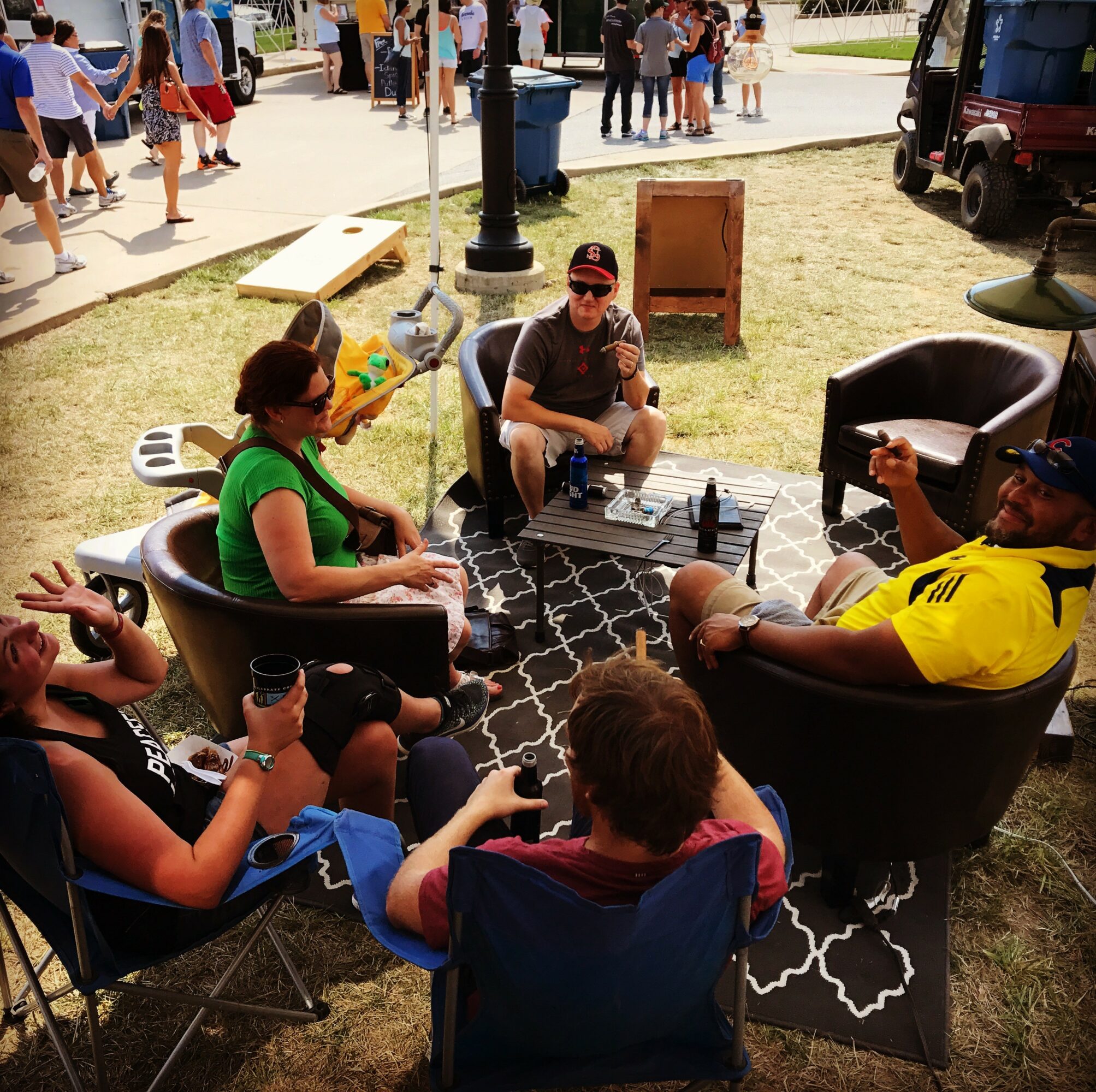 Group of five people sitting outdoors on chairs and a bench, talking and drinking, with a crowd in the background.